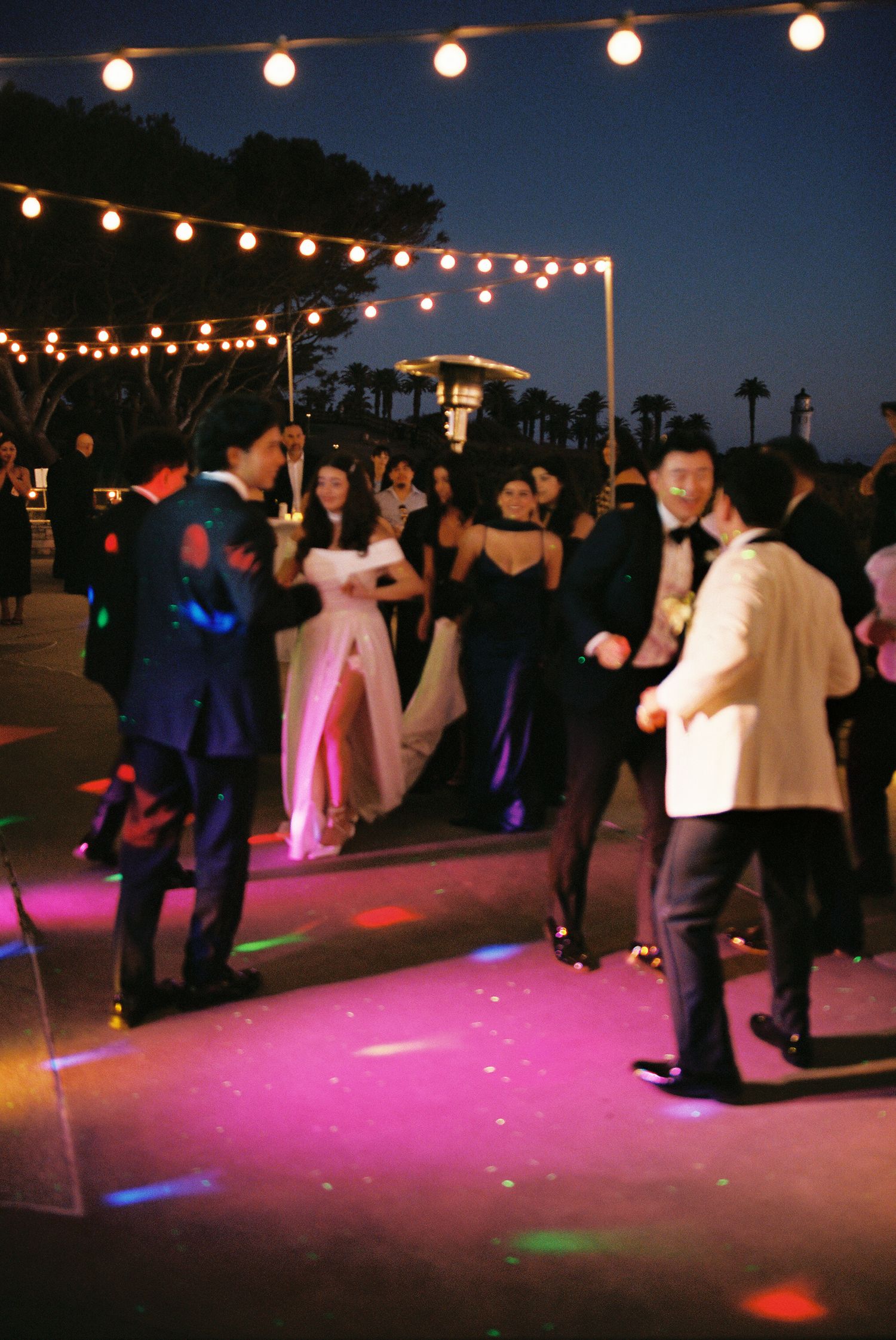 People dancing at an outdoor evening wedding reception under string lights with a pink-lit dance floor.