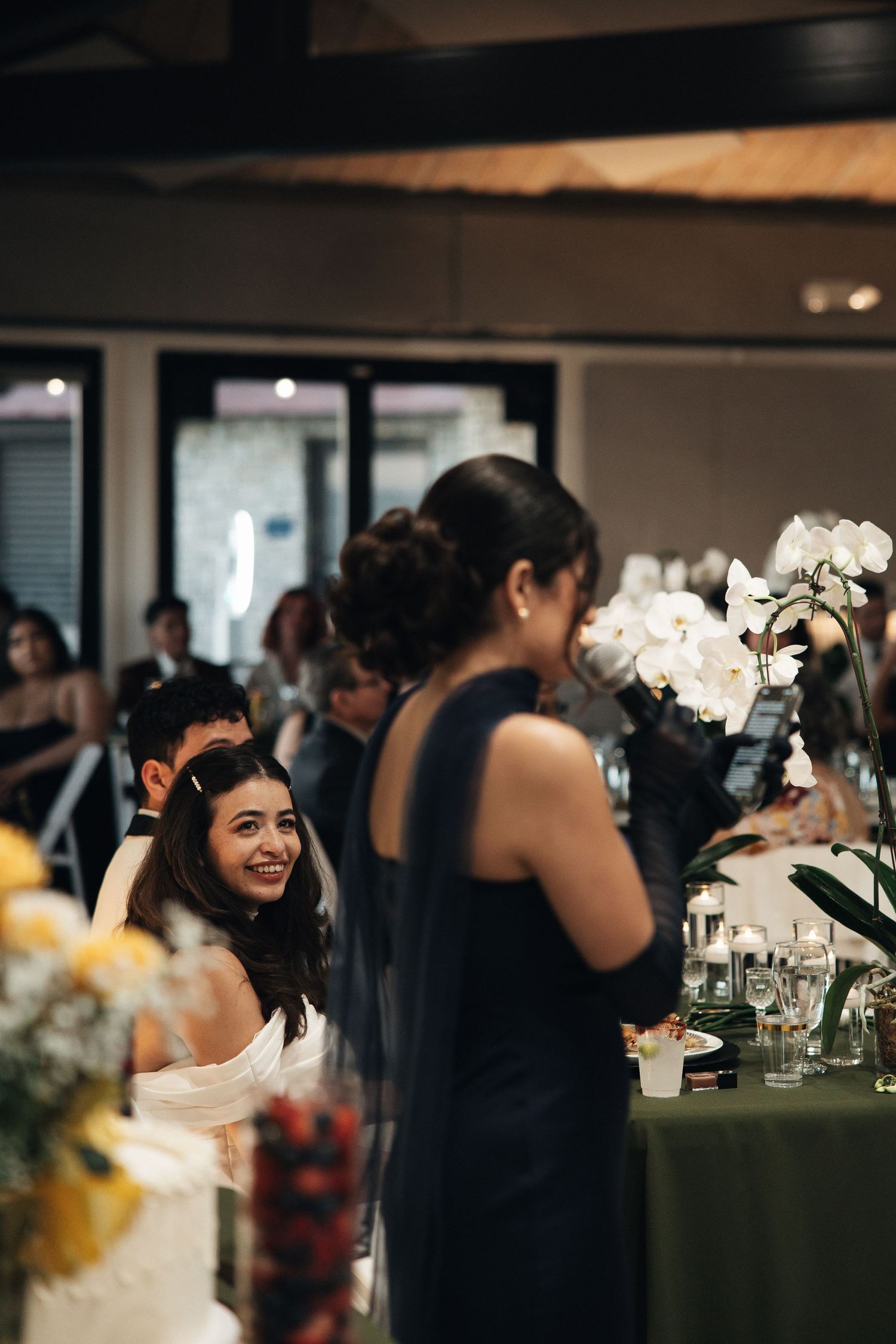 Wedding reception guests gather in an elegant indoor venue with white floral centerpieces and dark tablecloths.