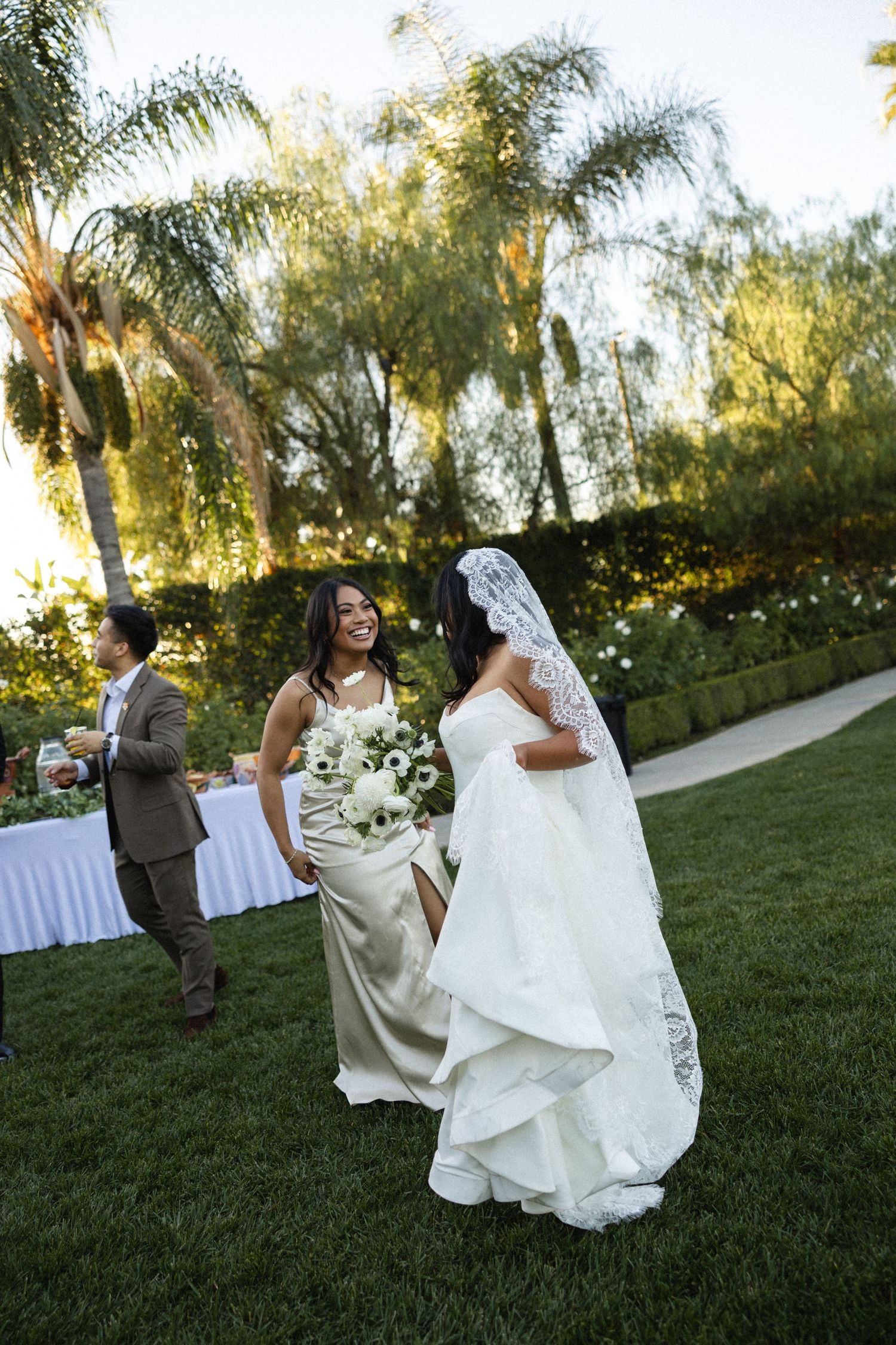 Wedding party members playfully dance and twirl on a sunlit lawn with palm trees in the background during golden hour.