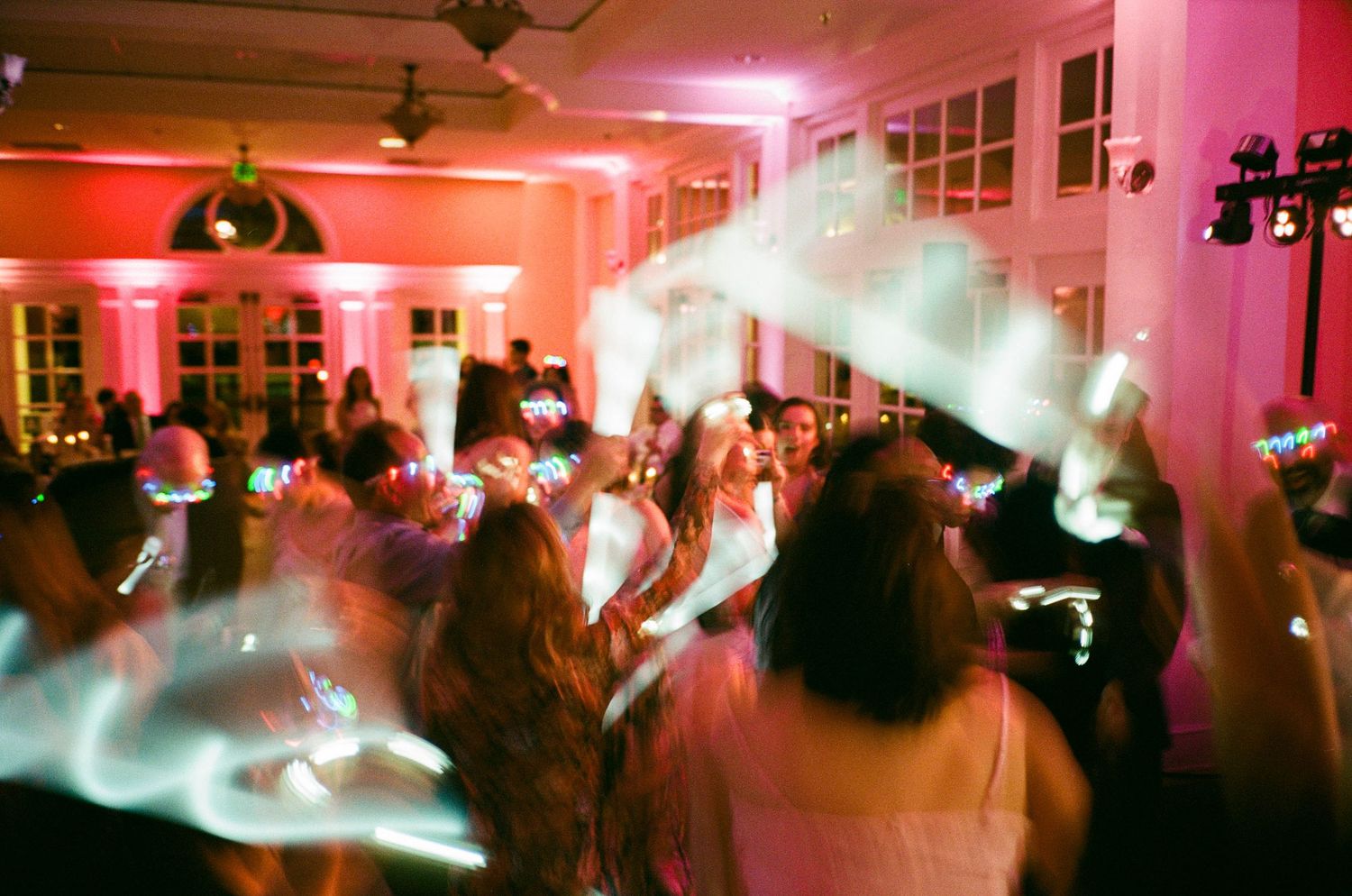 Energetic wedding reception dance floor with pink uplighting, glowing accessories, and motion-blurred guests celebrating at night.