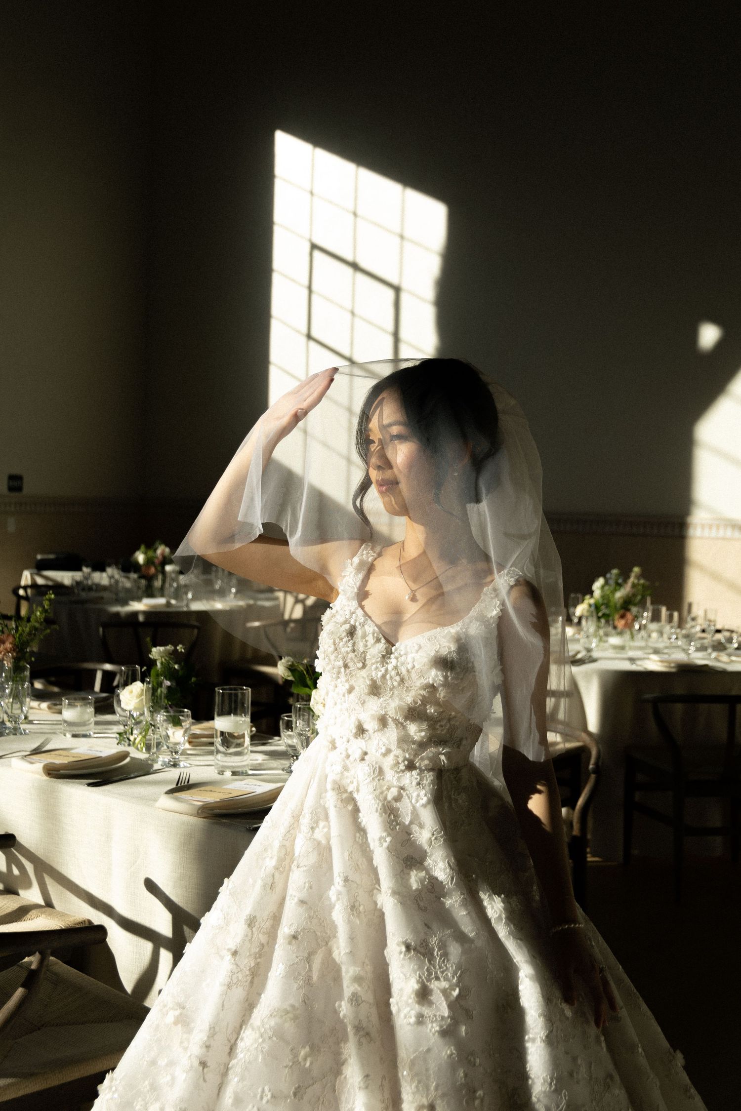 A bride in a white lace wedding dress stands in sunlit reception room with elegant table settings and floral arrangements.