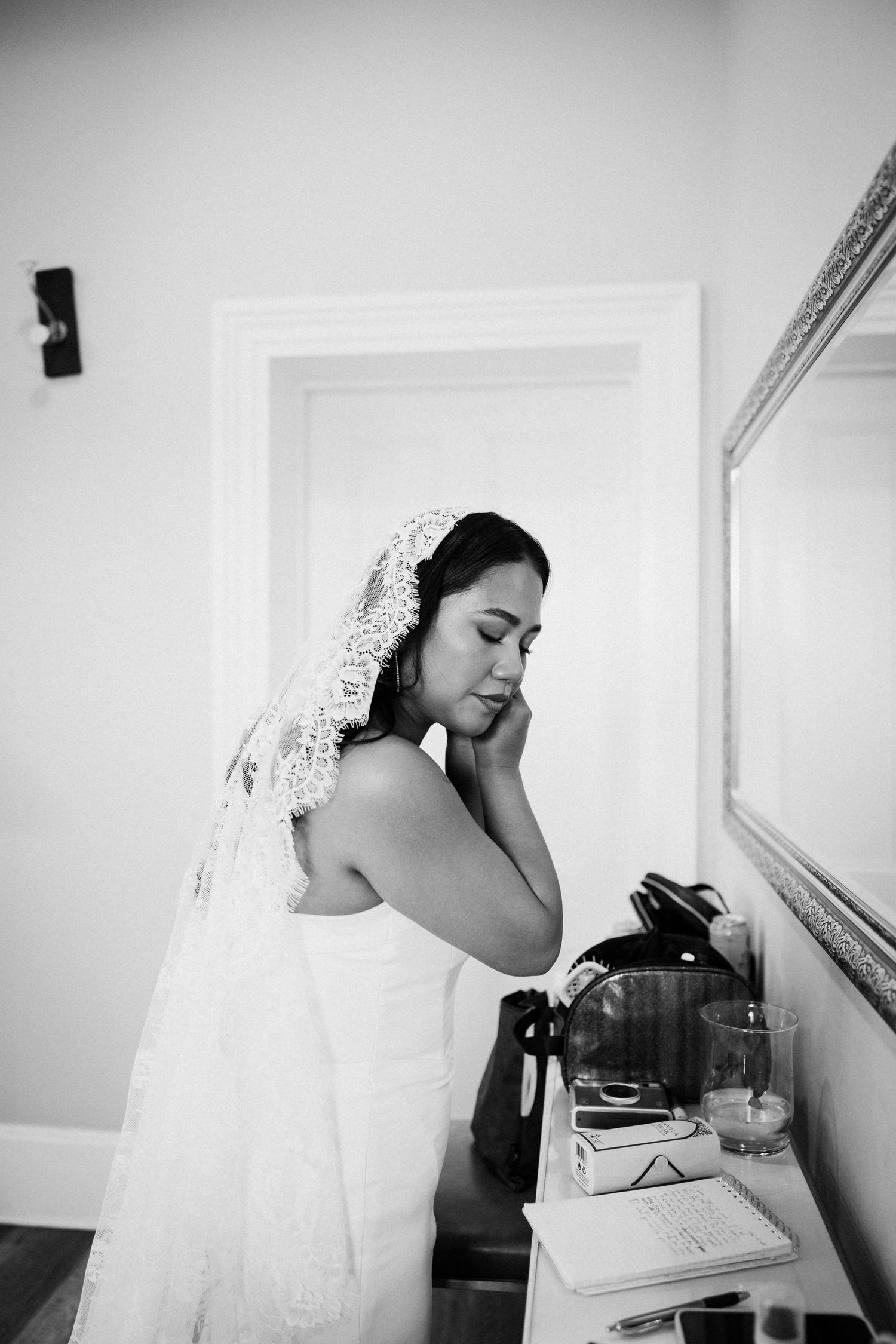 Black and white photo of a bride in a lace veil and white gown preparing at a vanity on her wedding day.