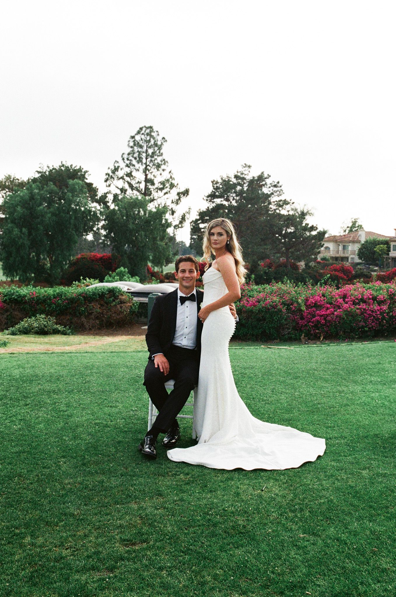 Bride in white gown and groom in black tuxedo pose on lush green lawn with colorful flowers and trees behind them.