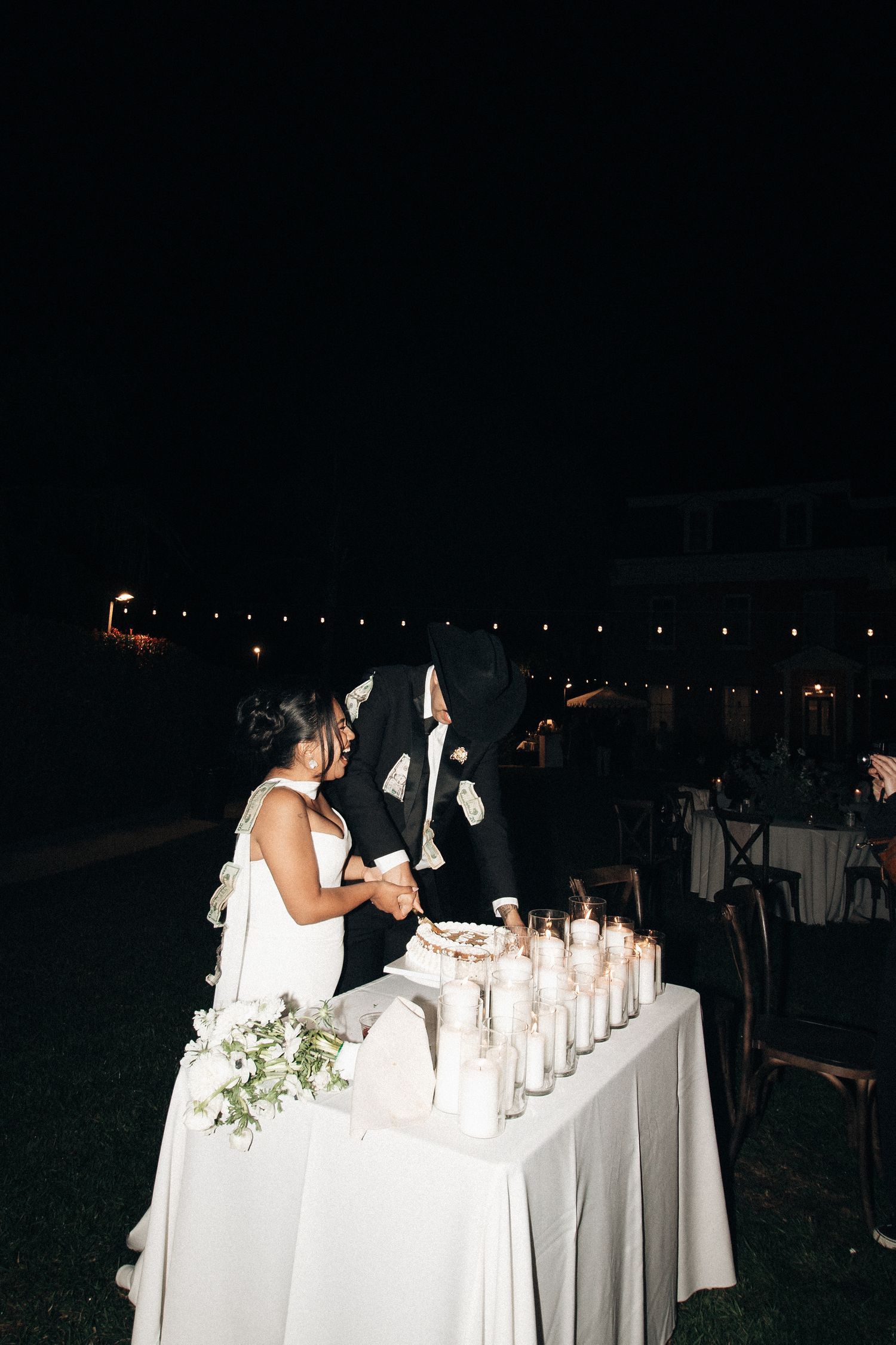 A series of glowing candles line a white-clothed table at an outdoor evening wedding reception.