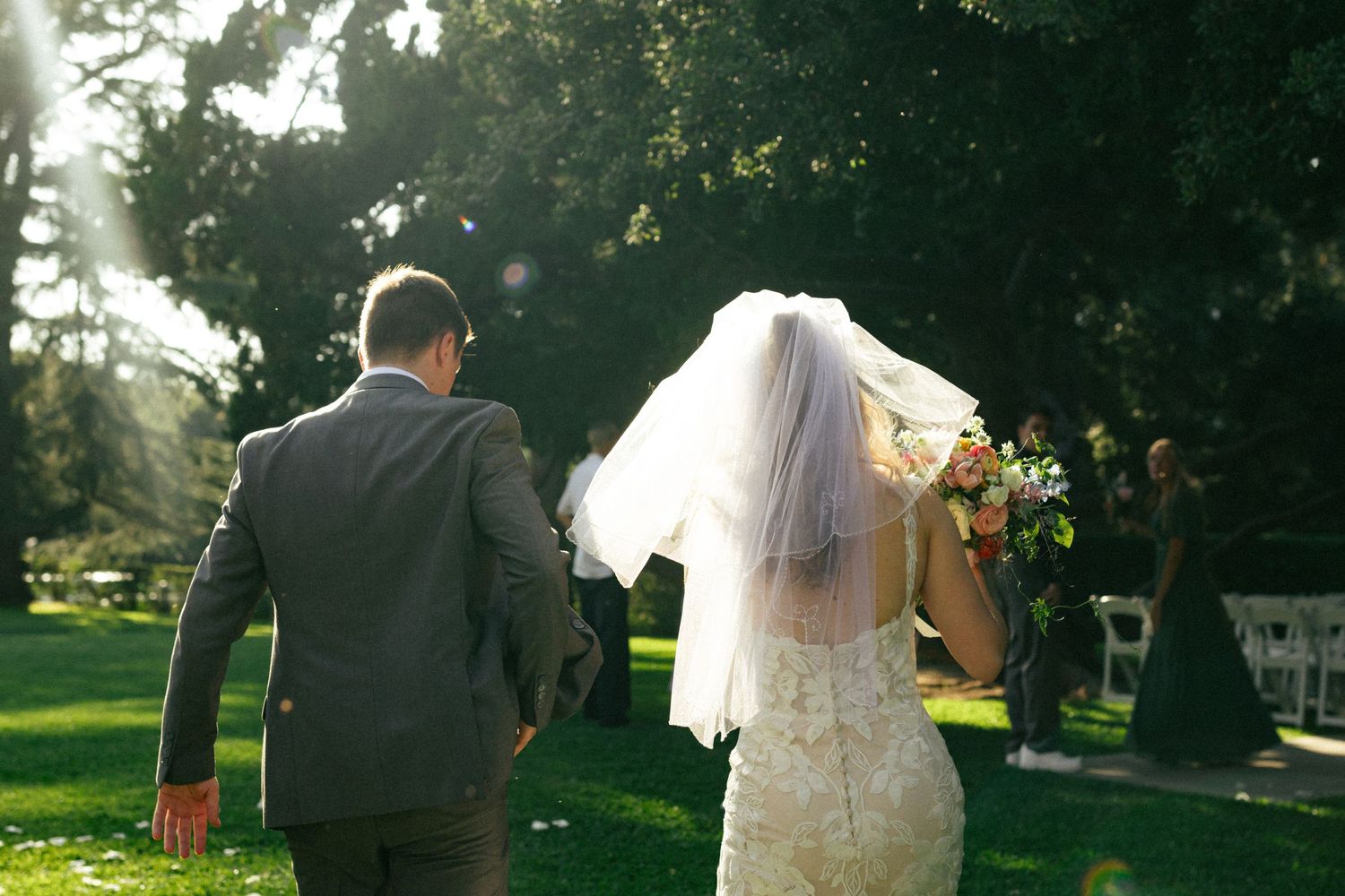 A newlywed couple walking in a sunlit garden park, with the bride in white dress and veil holding a bouquet.