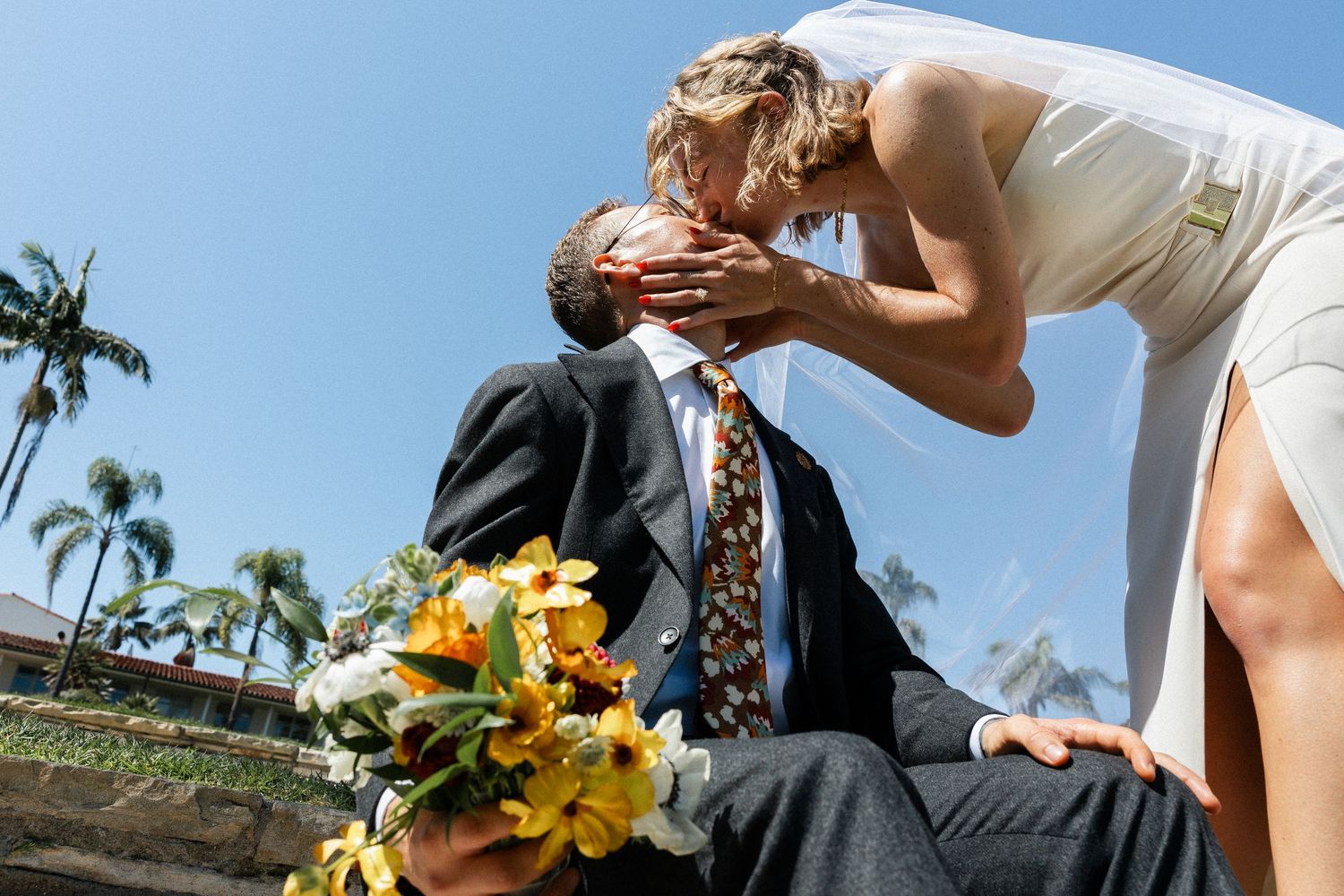Bride in white dress kissing groom in suit holding yellow flower bouquet outdoors on sunny wedding day.