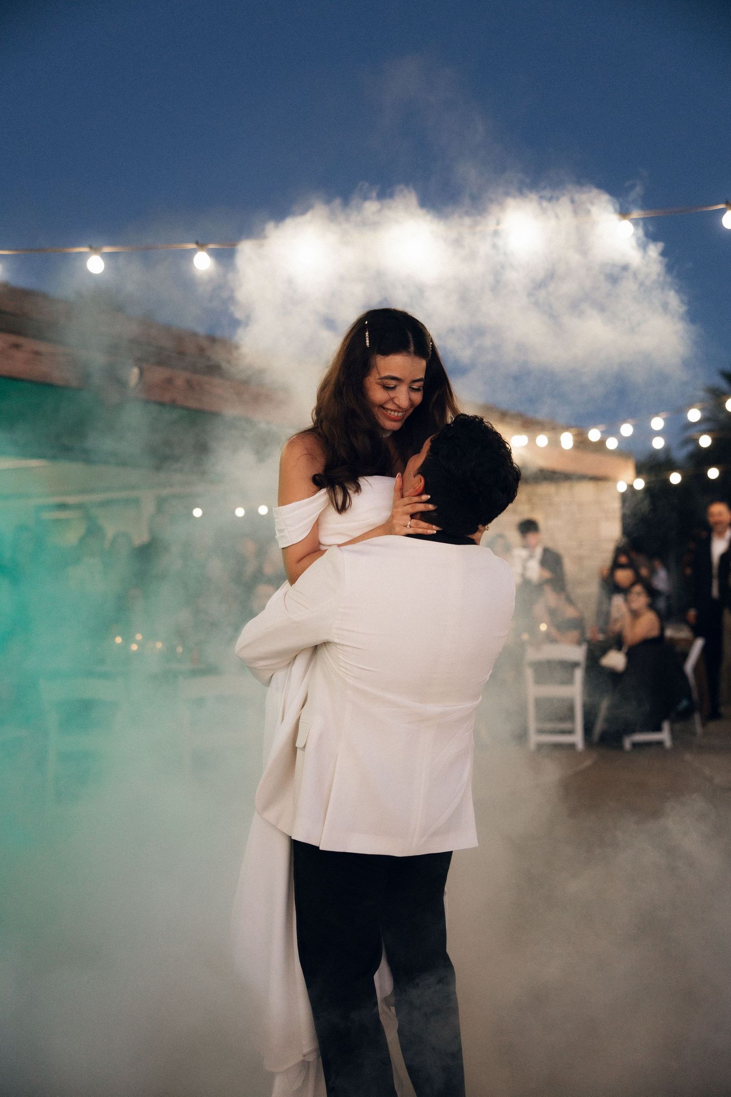 A couple shares a romantic dance surrounded by atmospheric fog and string lights at an outdoor evening reception.