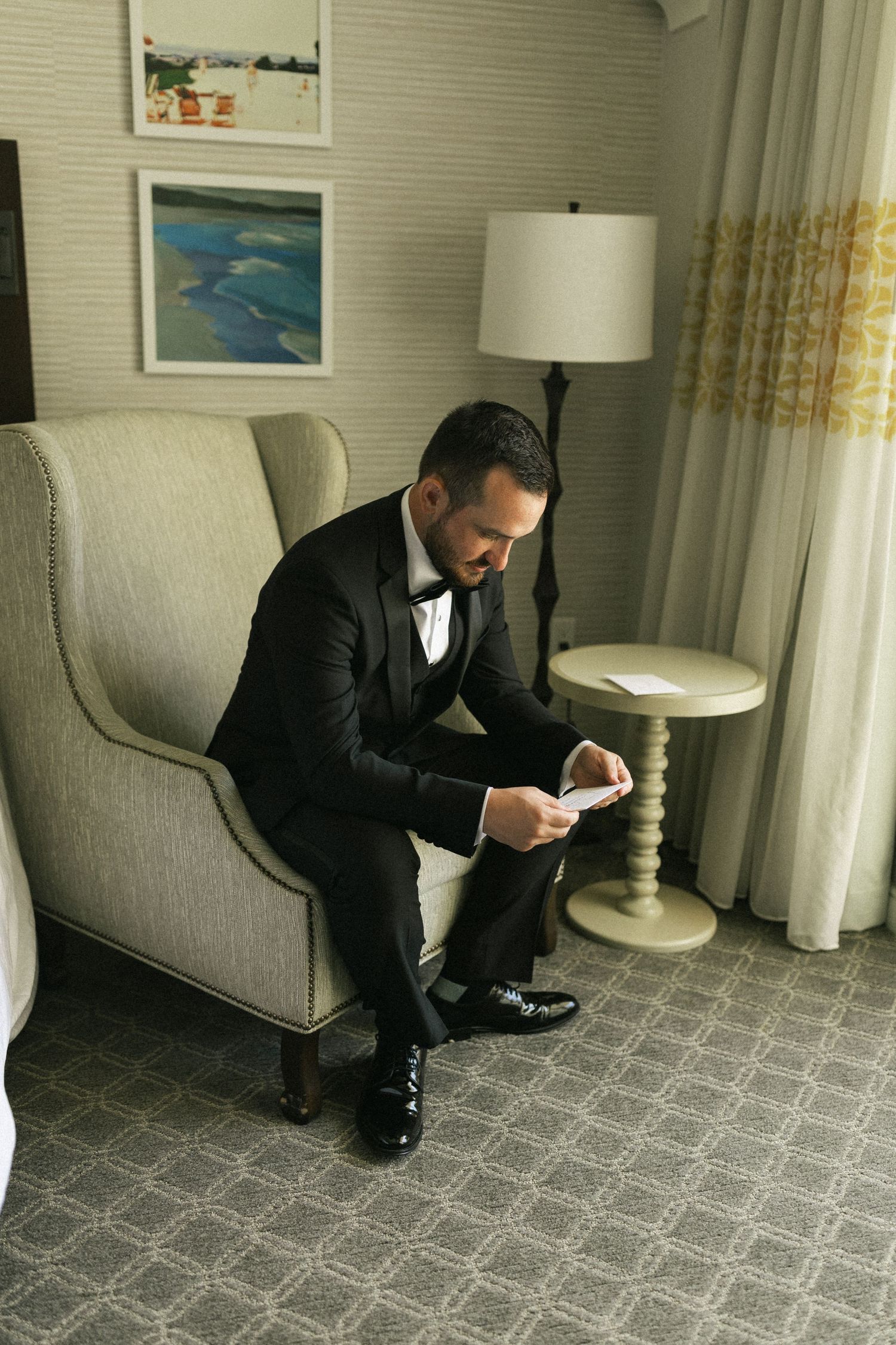 Someone in a black tuxedo sits in an upholstered gray armchair checking their phone in a hotel room setting.