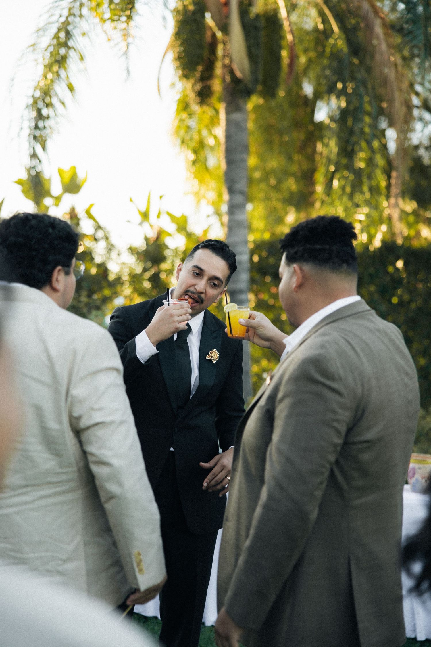 Three people in formal attire enjoying drinks and conversation at an outdoor event with trees in the background.