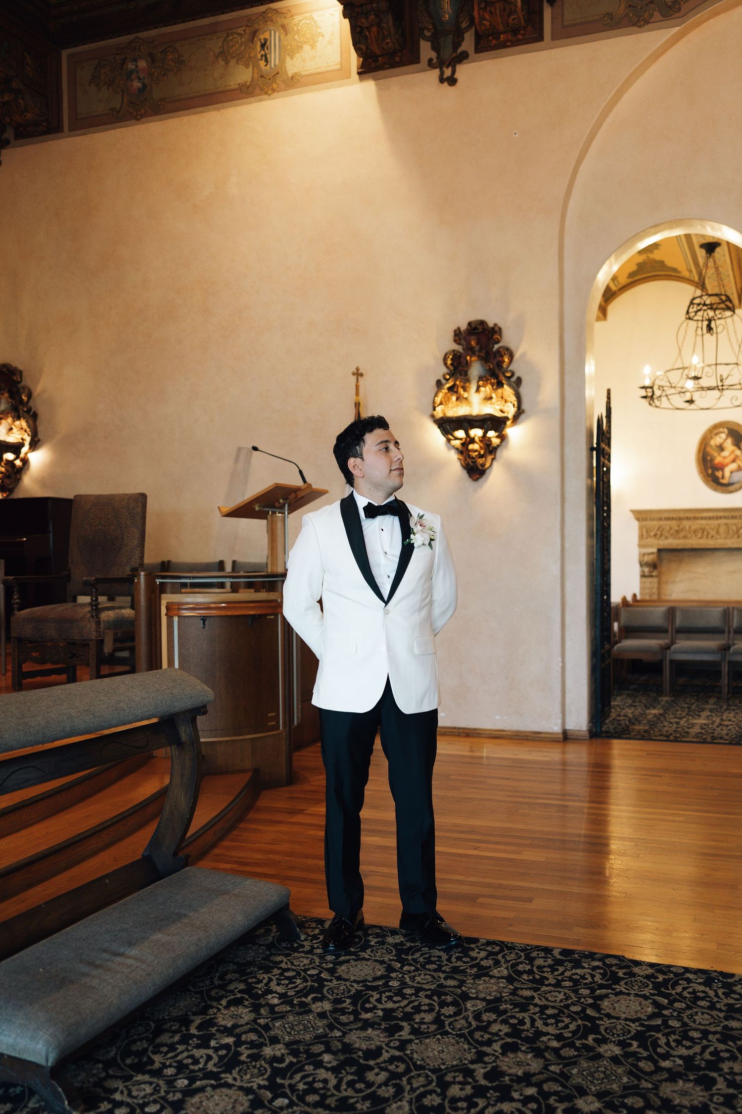A person in a white tuxedo jacket and black pants stands in a grand room with wooden floors and ornate decor.