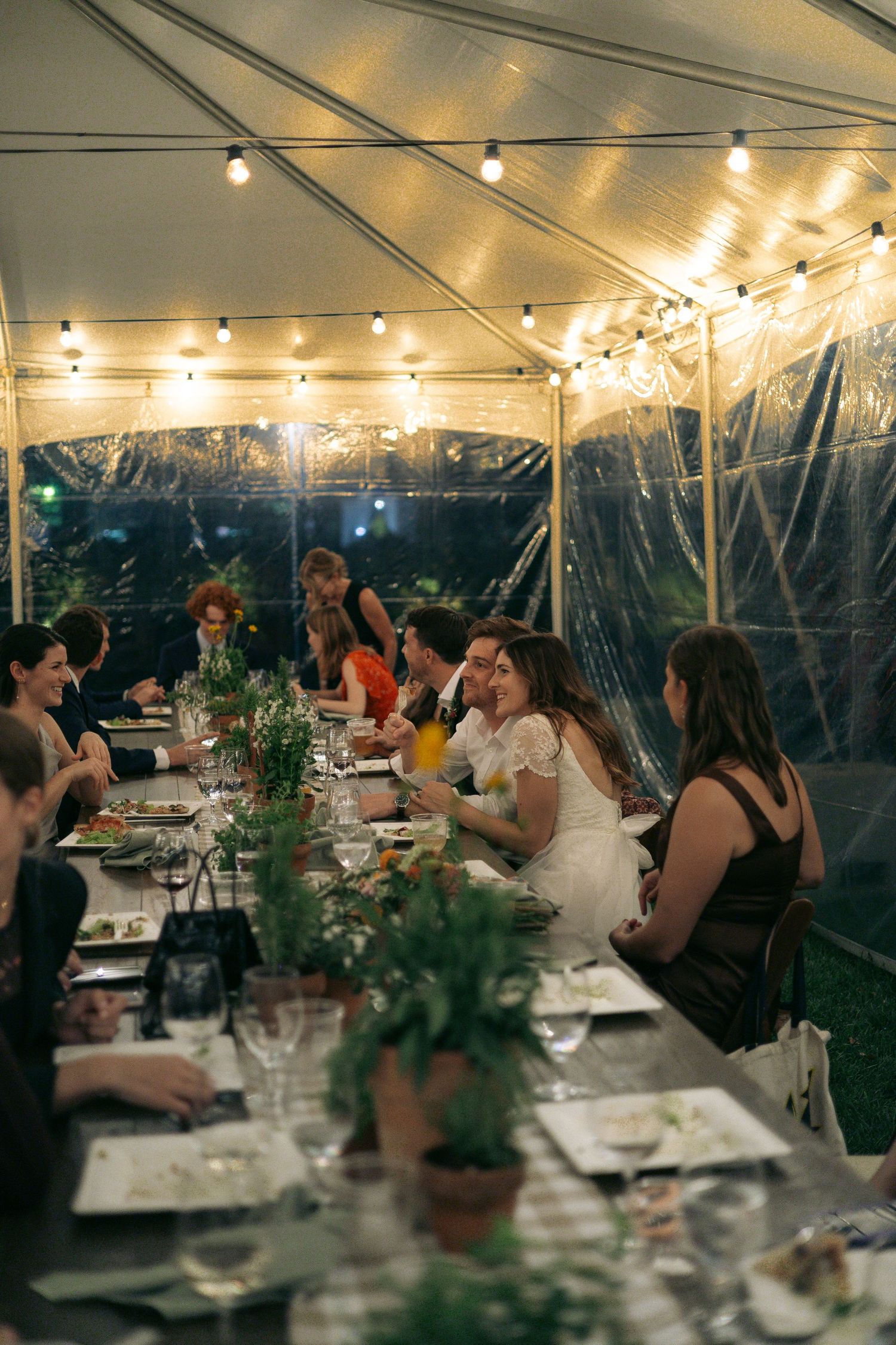 Guests dine at an outdoor evening event under a tent with string lights and greenery centerpieces.