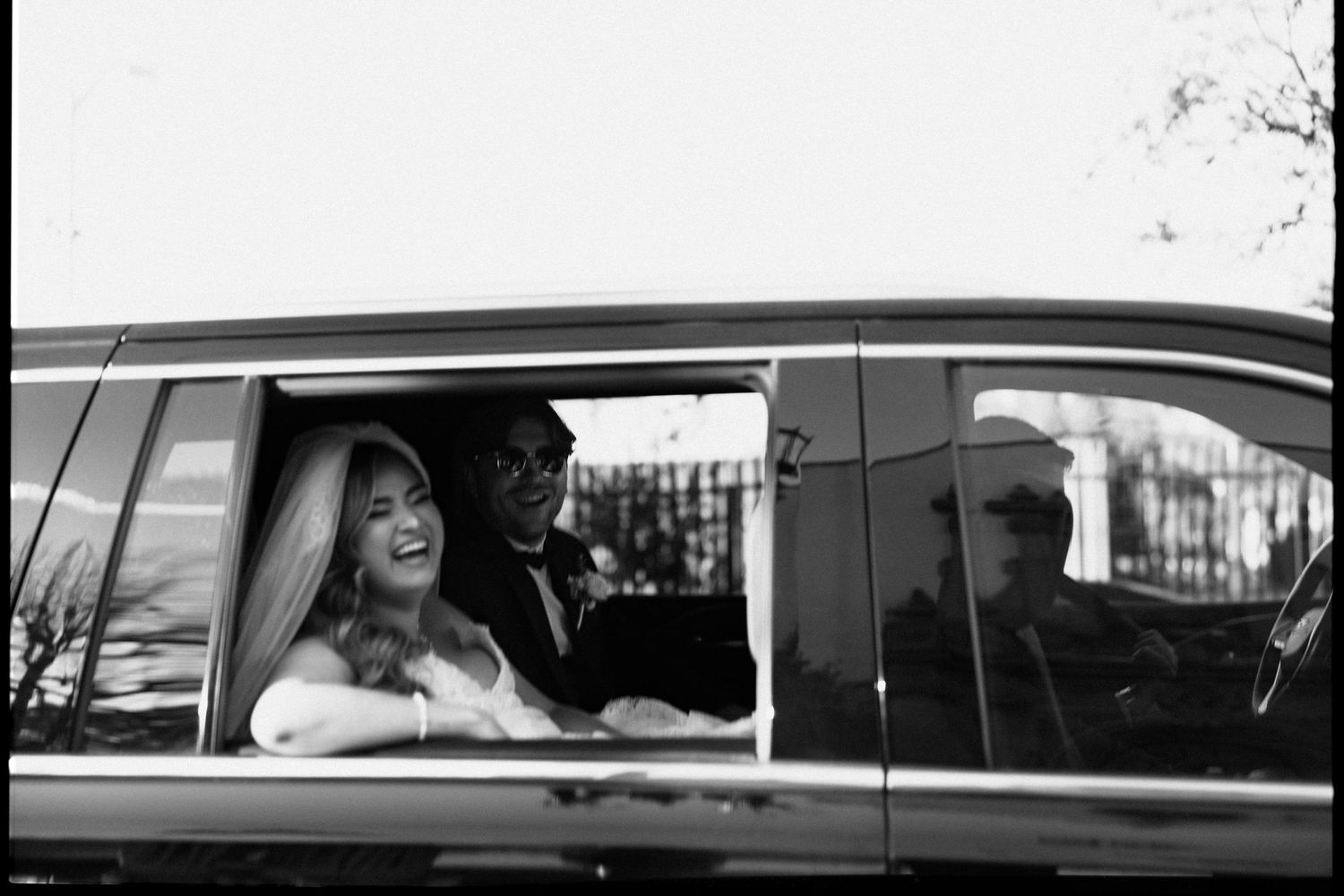 Black and white photo of a smiling bride in a wedding veil sitting in a car with groom on their wedding day.