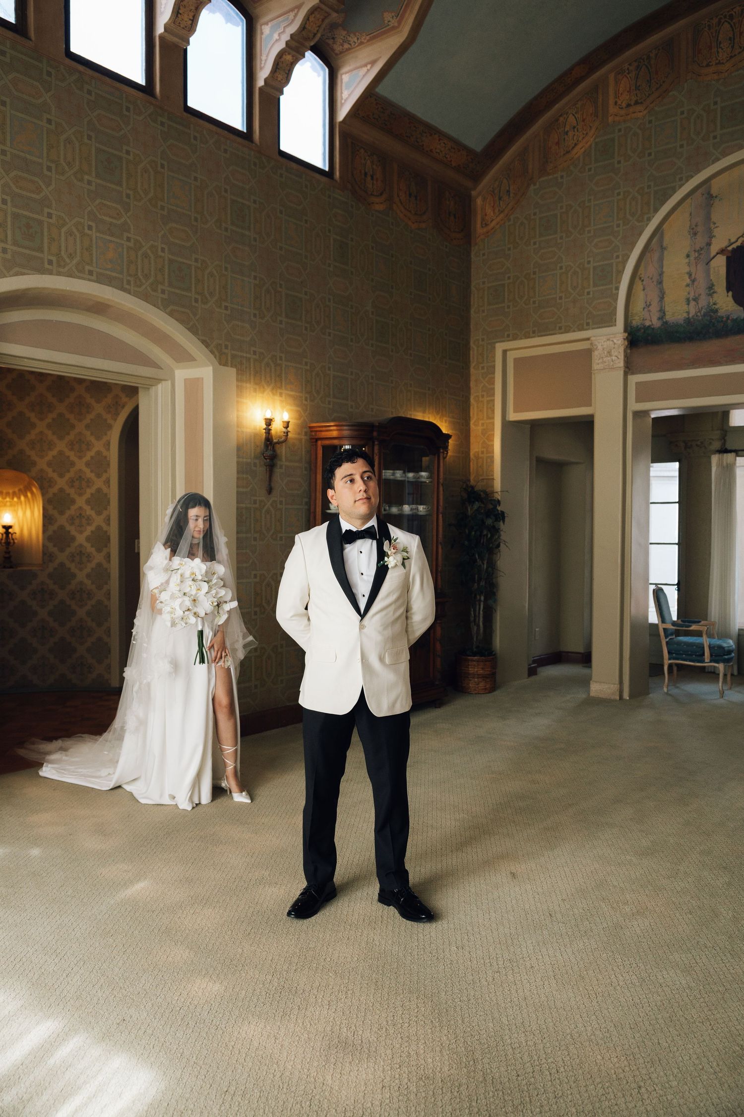 A couple poses in an elegant venue with ornate wallpaper and arched doorways, the groom in a white tuxedo jacket.