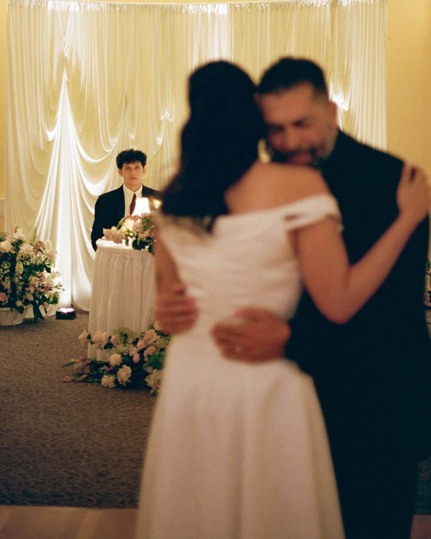 Couple sharing first dance at wedding reception, with officiant standing near floral arrangements and draped backdrop.