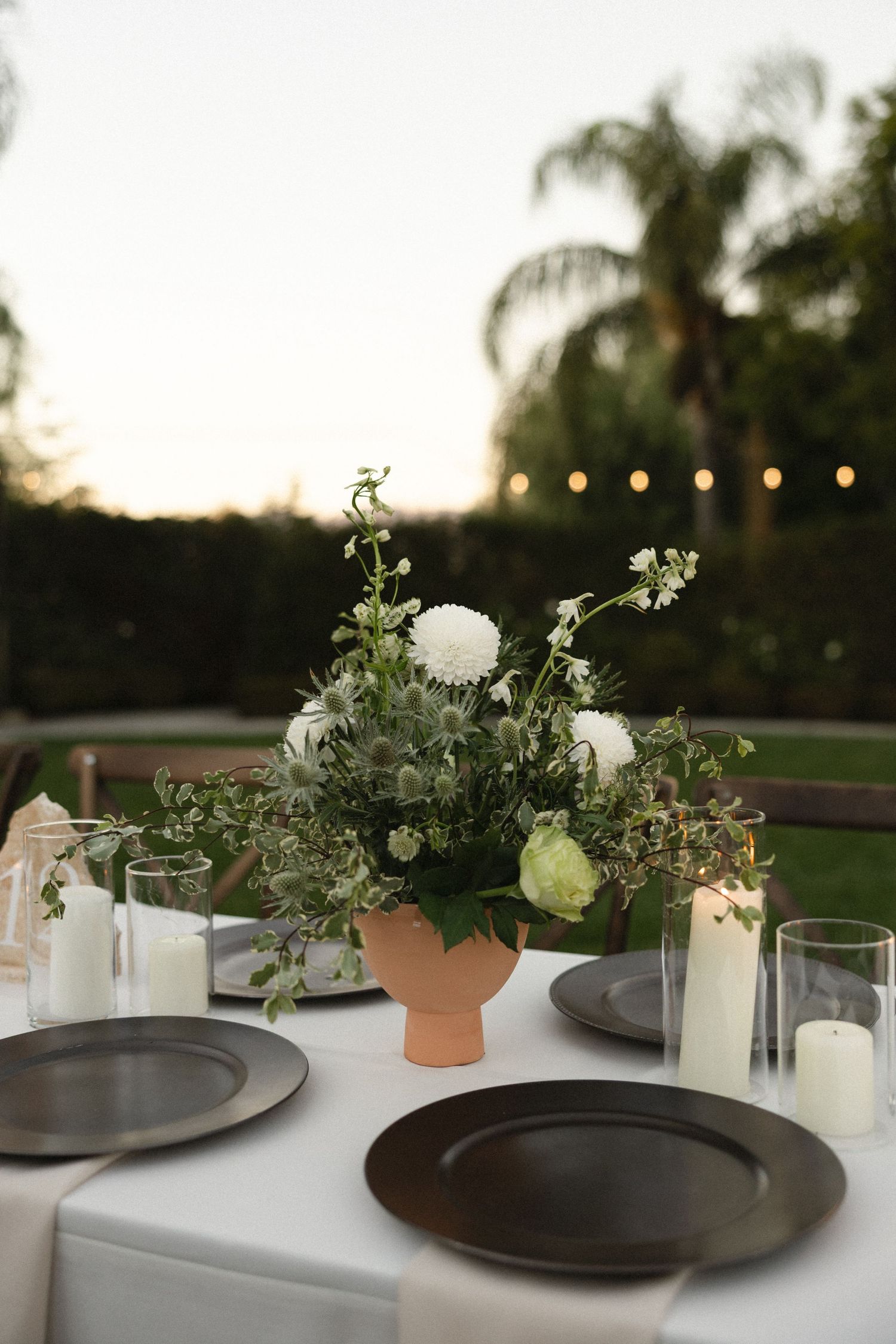 Elegant outdoor table setting with black plates, white linens, and delicate floral centerpieces at sunset.
