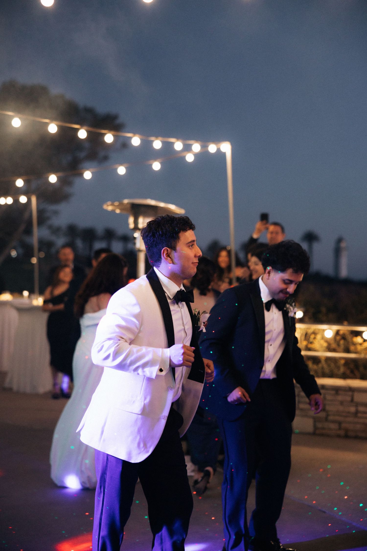 Wedding guests dance under twinkling string lights at an outdoor evening reception.