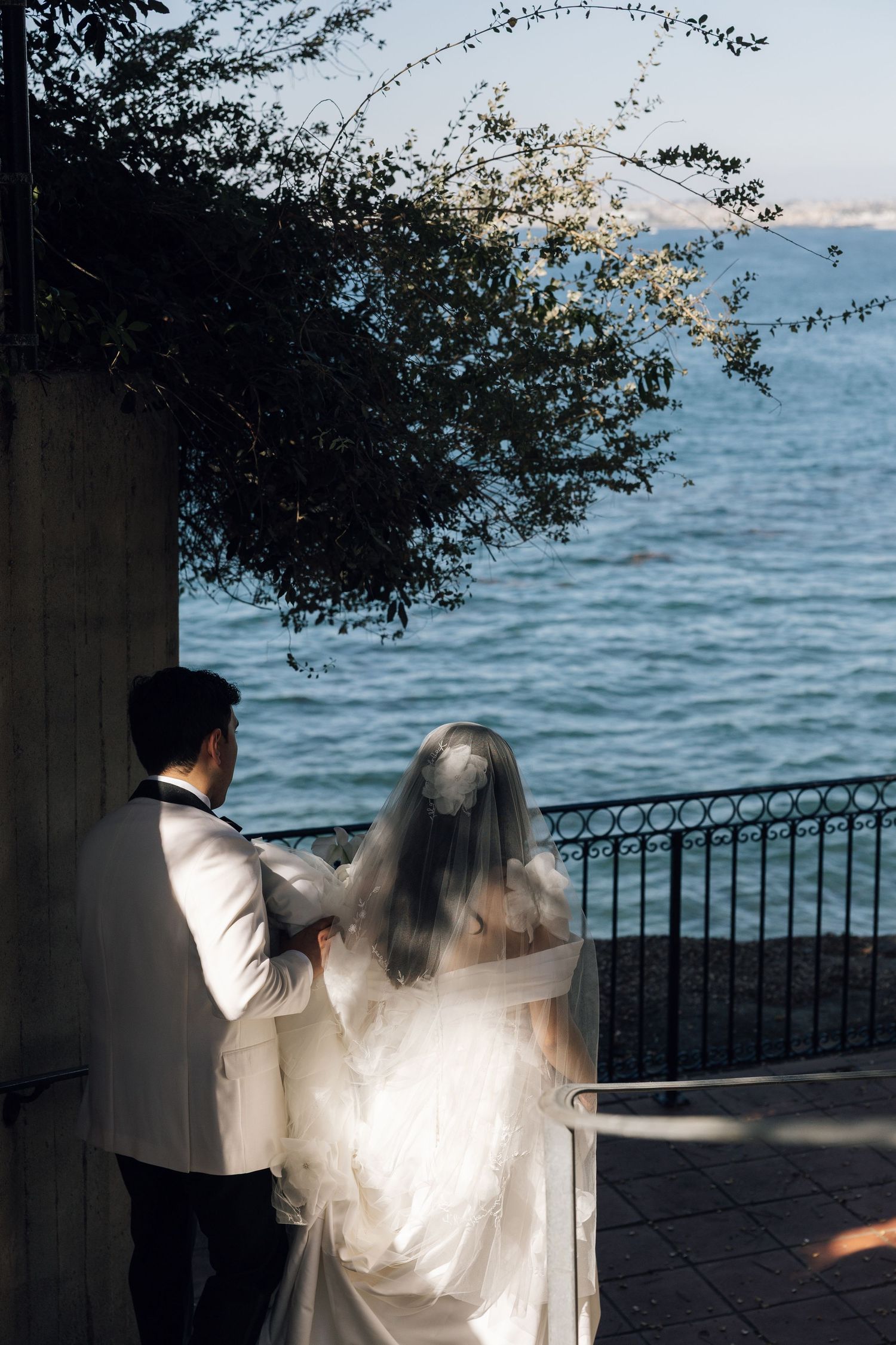 A couple in wedding attire stands by a lakeside railing at sunset with tree branches framing the romantic scene.