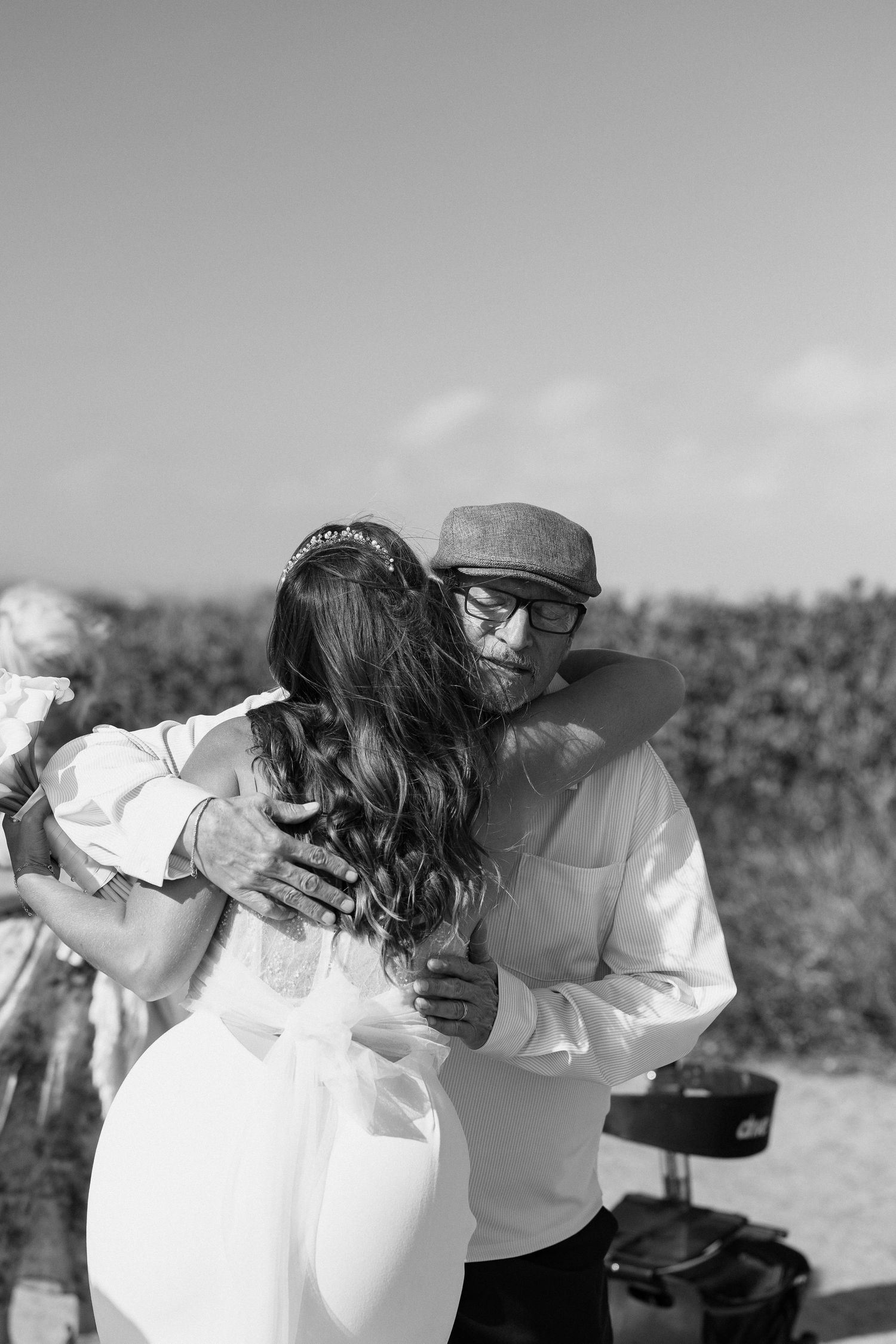 Black and white photo of a bearded groom in a cap embracing his bride in a white dress outdoors.