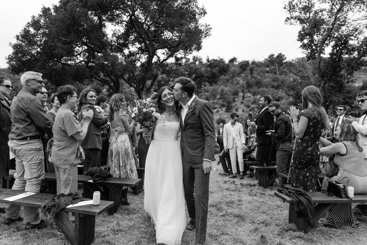 A black and white outdoor wedding ceremony shows a couple sharing a moment surrounded by guests standing on logs.