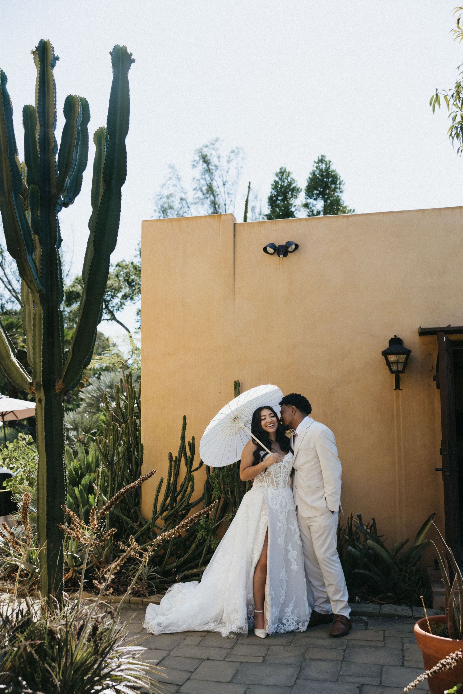 A couple poses in front of a beige wall with tall desert cactus plants at an outdoor wedding venue. Micro wedding, intentional celebrations