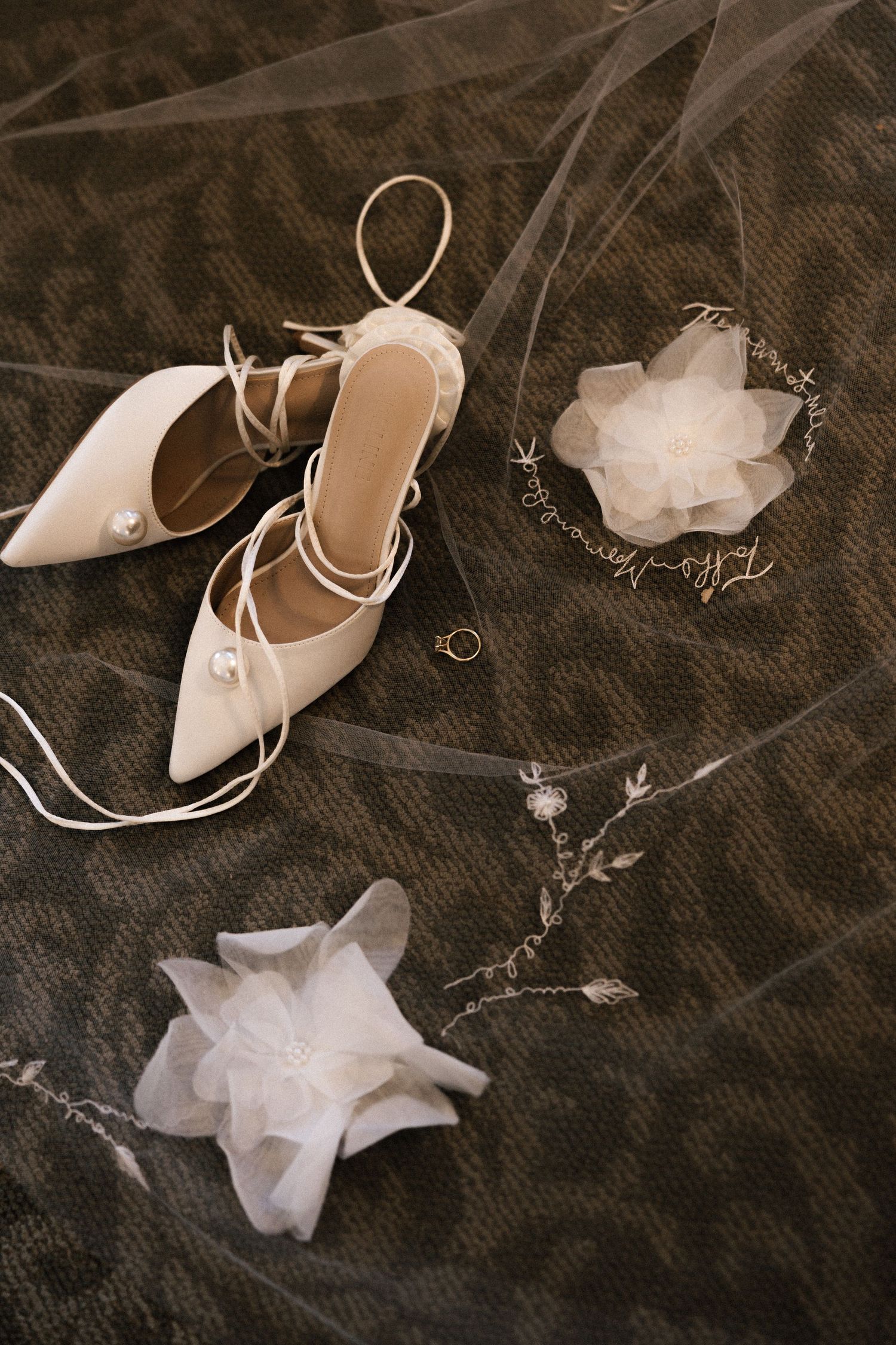 White bridal accessories including delicate shoes, veil, and jewelry arranged on dark textured fabric.