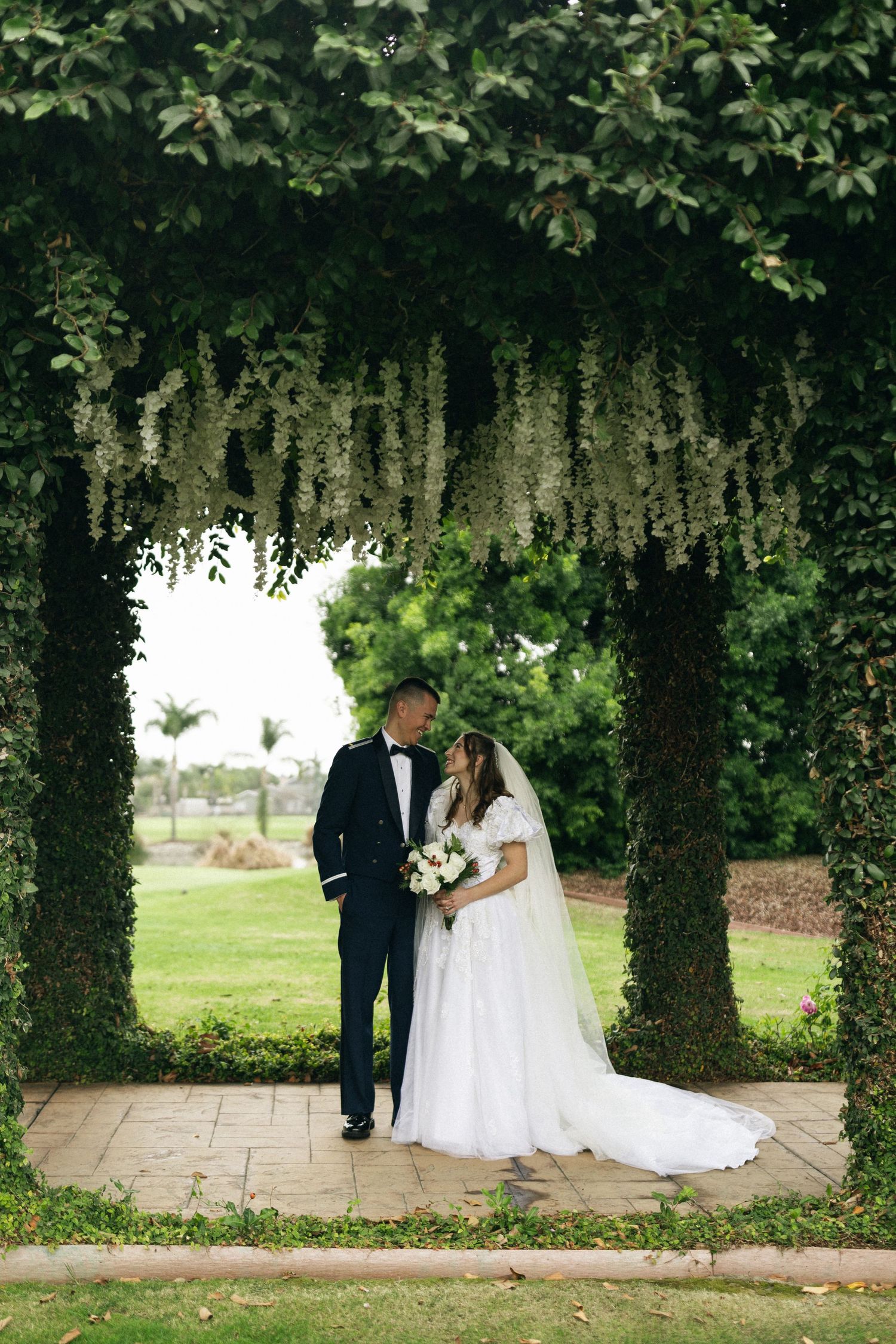 A romantic wedding couple stands beneath an archway covered in cascading vines and greenery on a stone path.