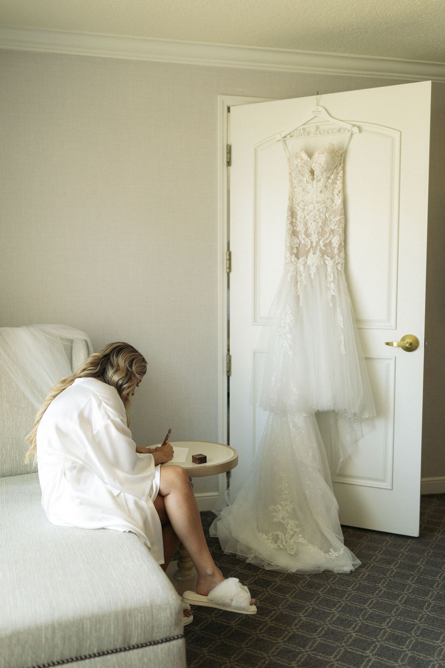 A beautiful white lace wedding dress hangs on a door while someone in a robe sits on a couch writing vows. Documentary/ photojournalist phot