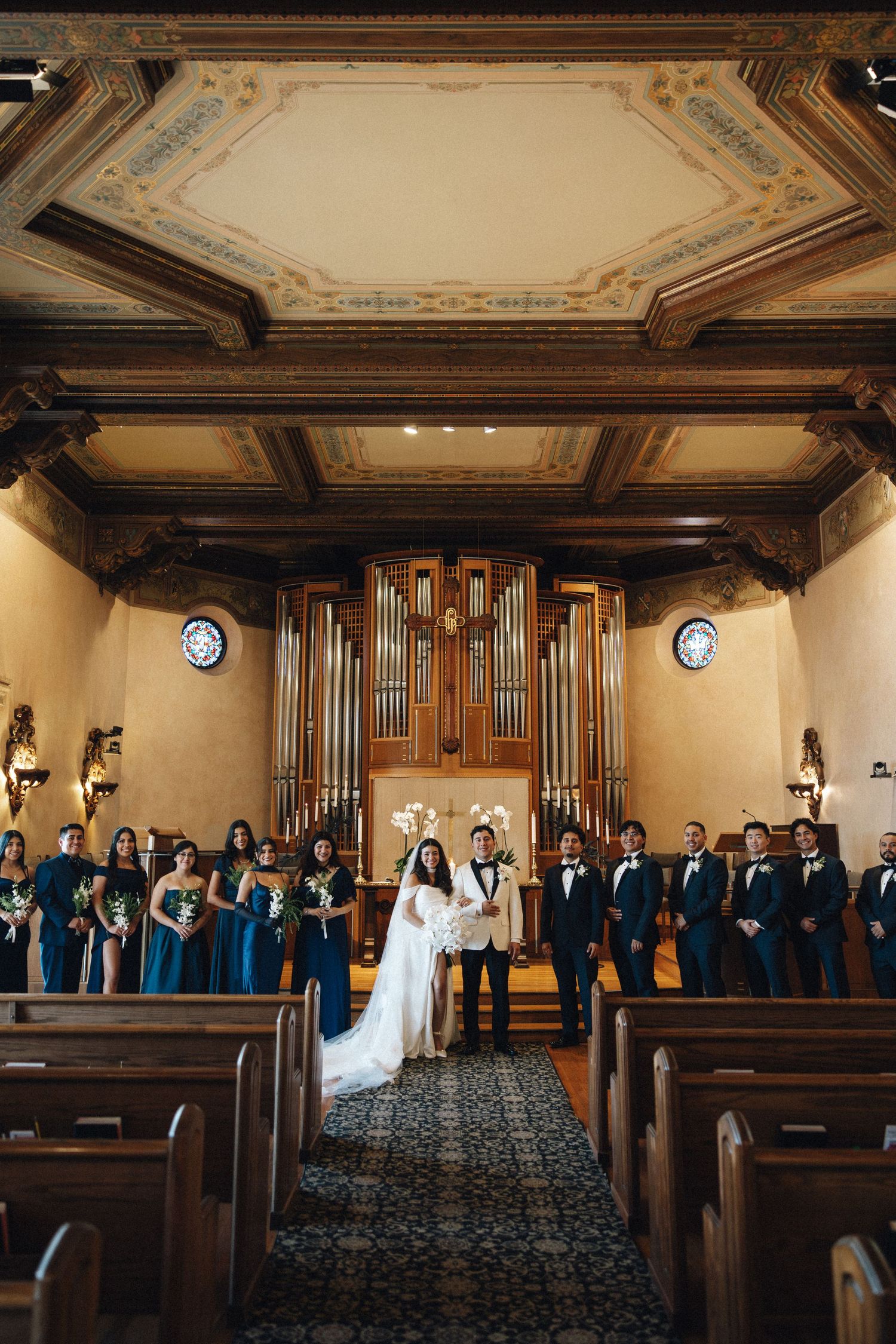Wedding party stands in front of grand church organ pipes with ornate ceiling and wooden pews in traditional chapel.