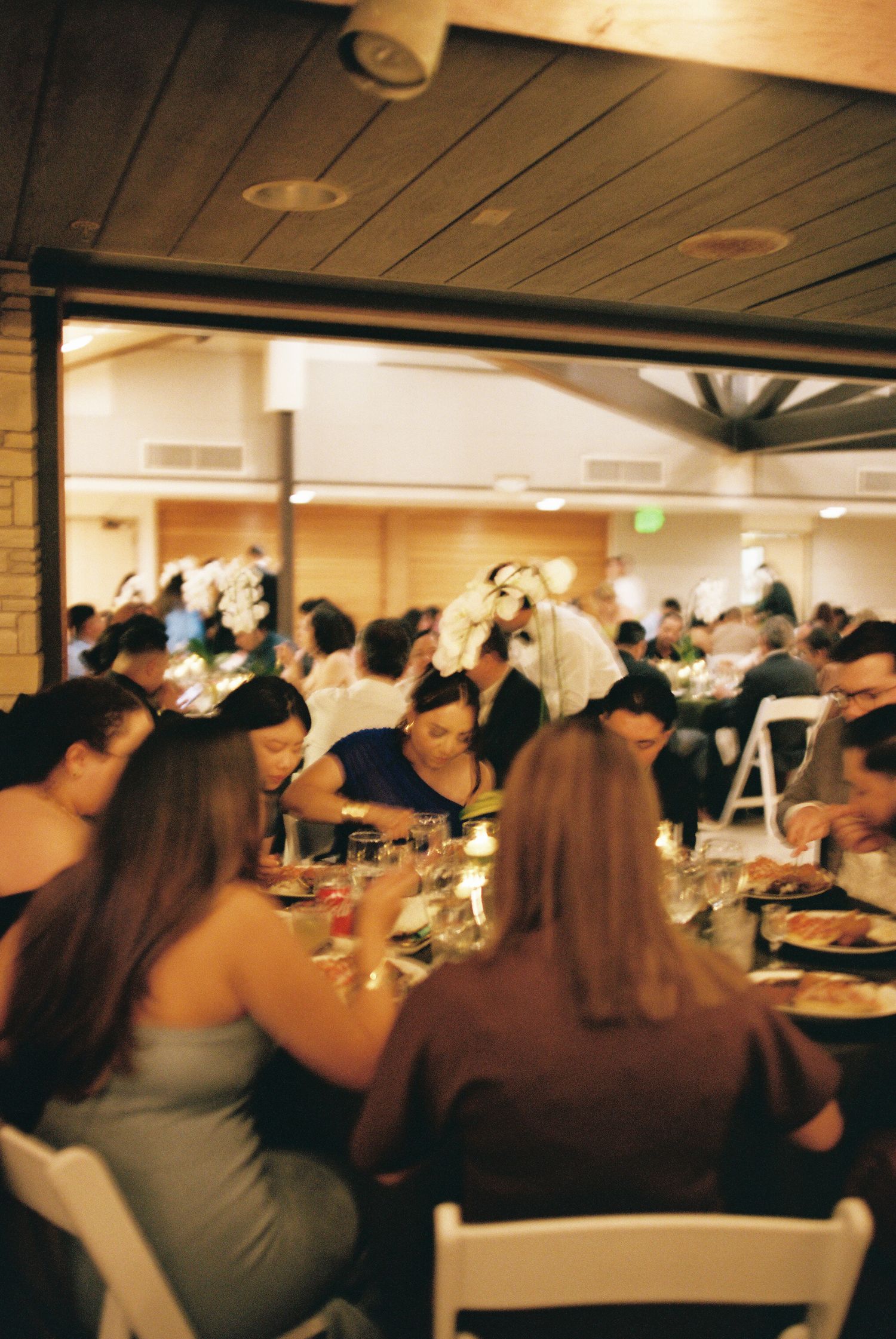Crowded indoor dining event with people seated at tables in a large room under wooden ceiling beams.