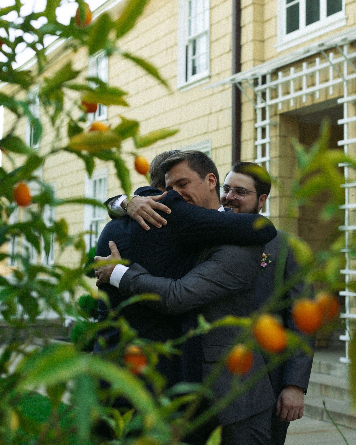 A warm embrace between two individuals in suits takes place in a garden with citrus trees in the foreground.