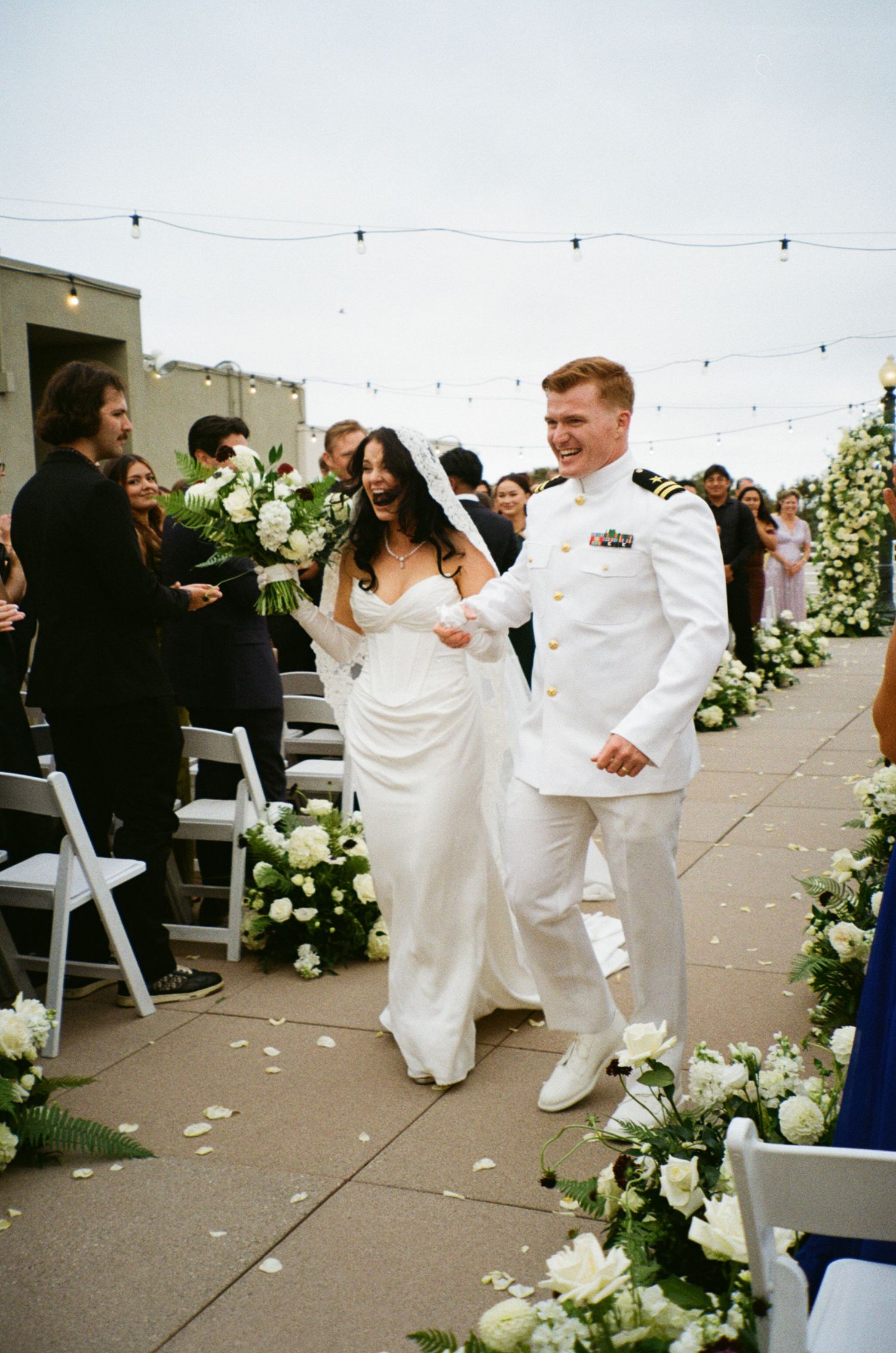 Bride and groom in white Navy uniform walk joyfully down outdoor aisle lined with white floral arrangements.