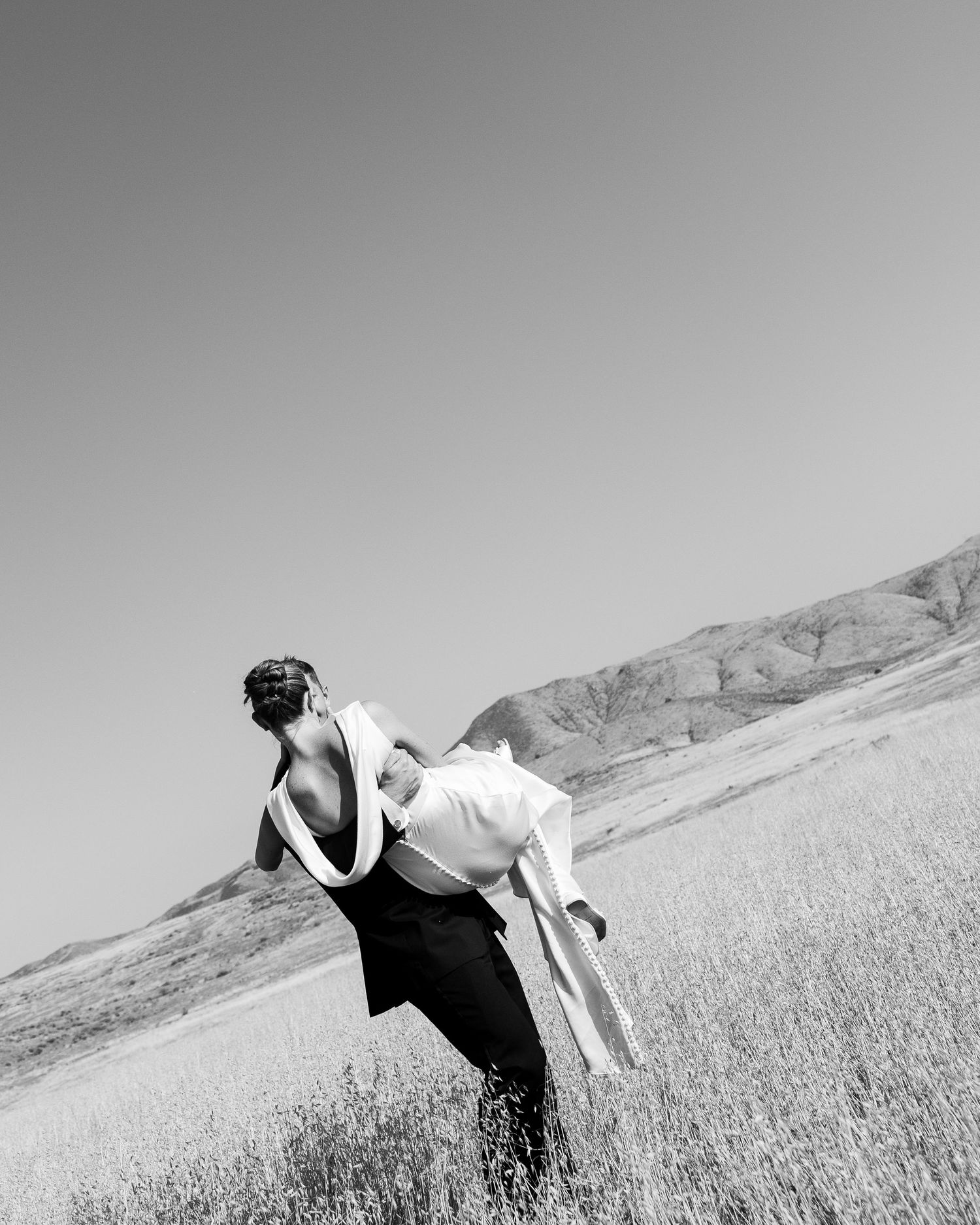 Black and white photo of a person carrying another through a grassy field with hills and clear sky in background.