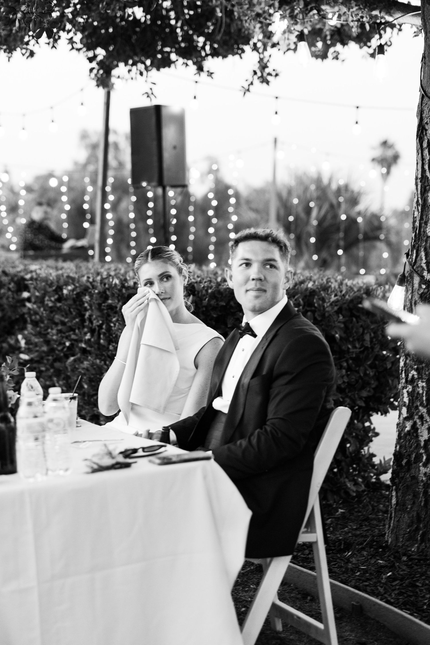 Black and white photo of bride wiping tears and groom smiling at outdoor wedding reception with string lights.