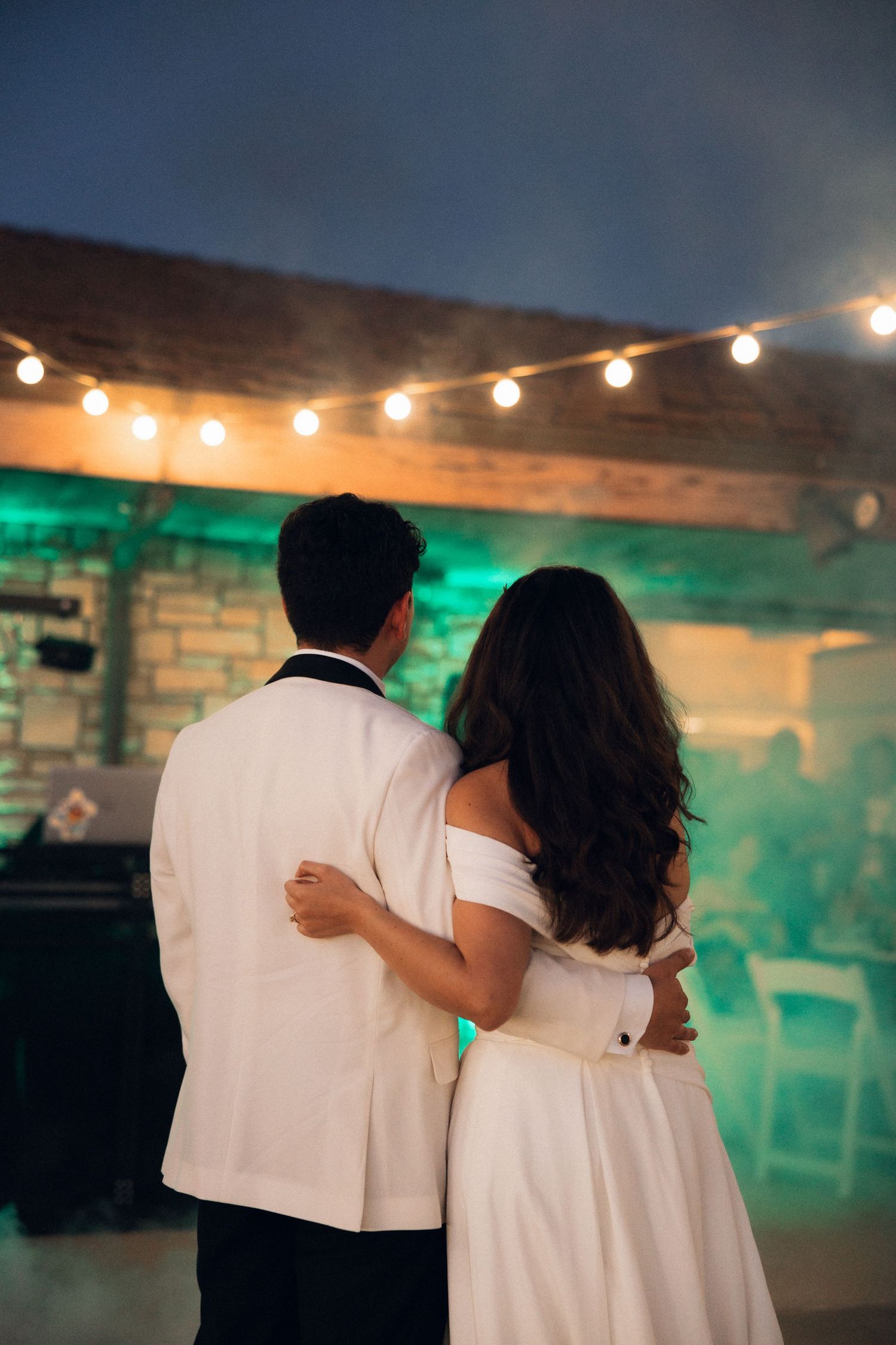 A couple in white outfits dances together under string lights at an evening reception, creating a romantic silhouette.