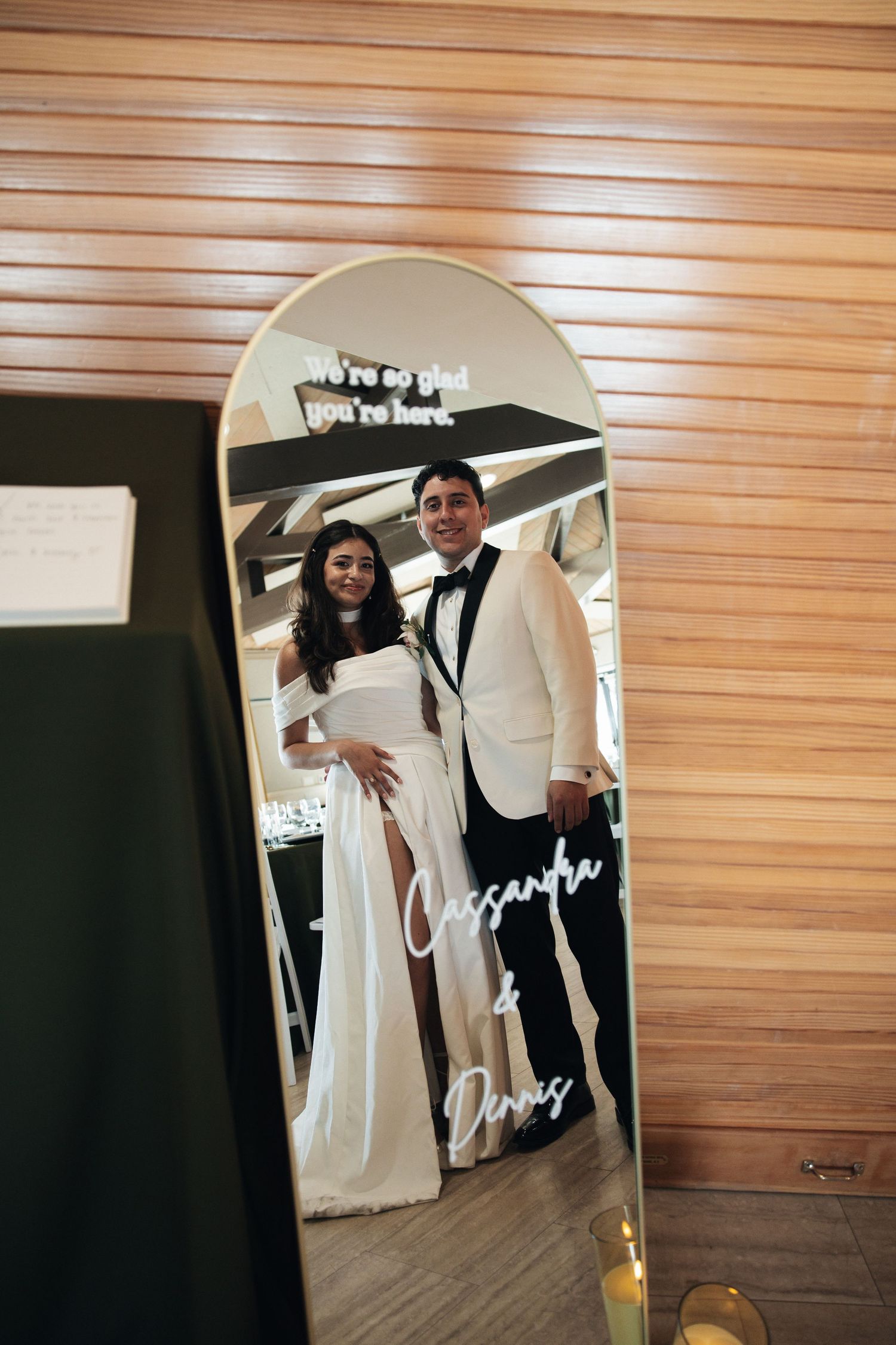 A wedding dress and suit are reflected in a mirror with a handwritten signature visible on the glass surface.