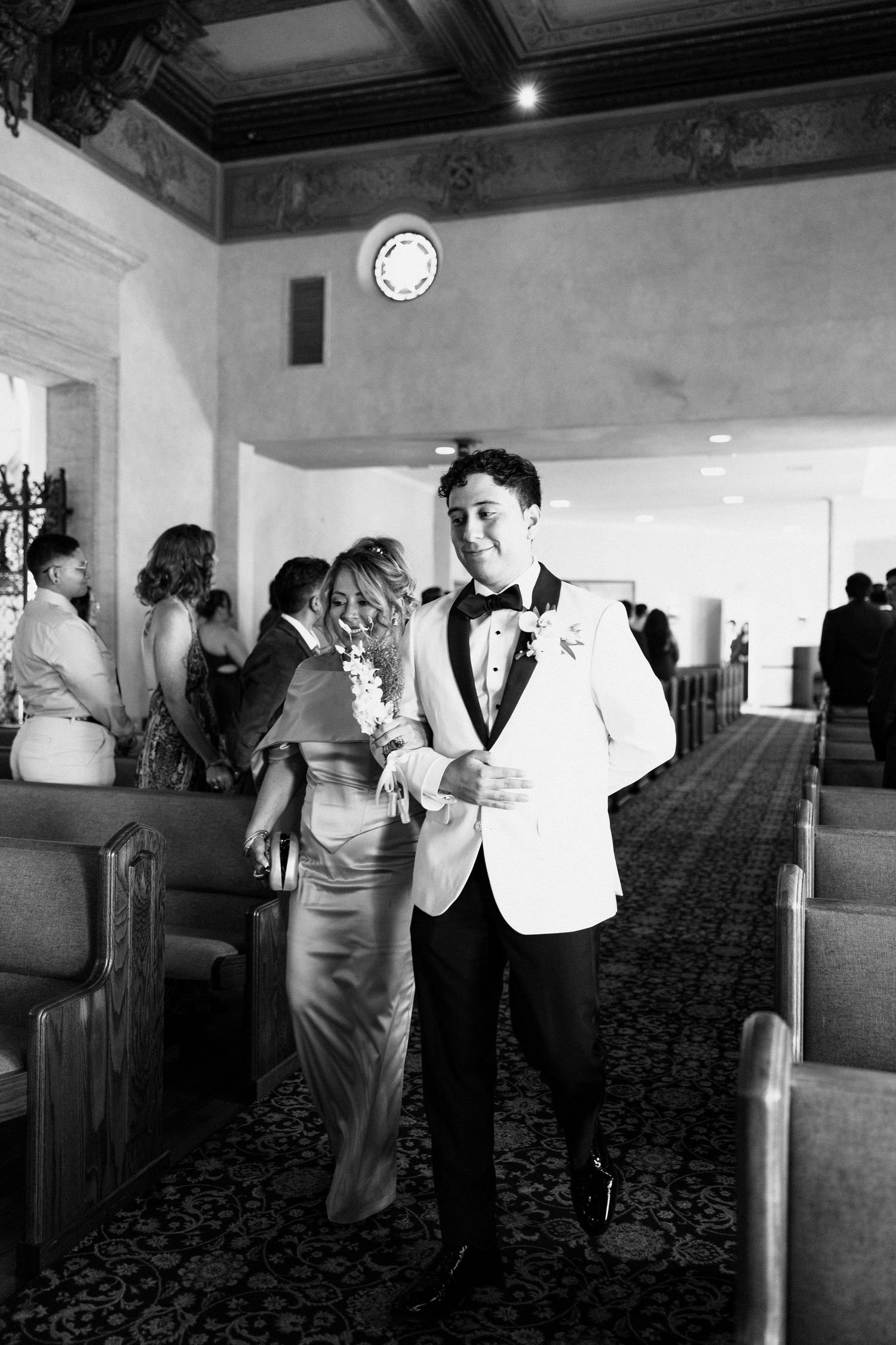 A couple in formal attire walks down an aisle at an elegant venue, with the groom wearing a white tuxedo jacket.