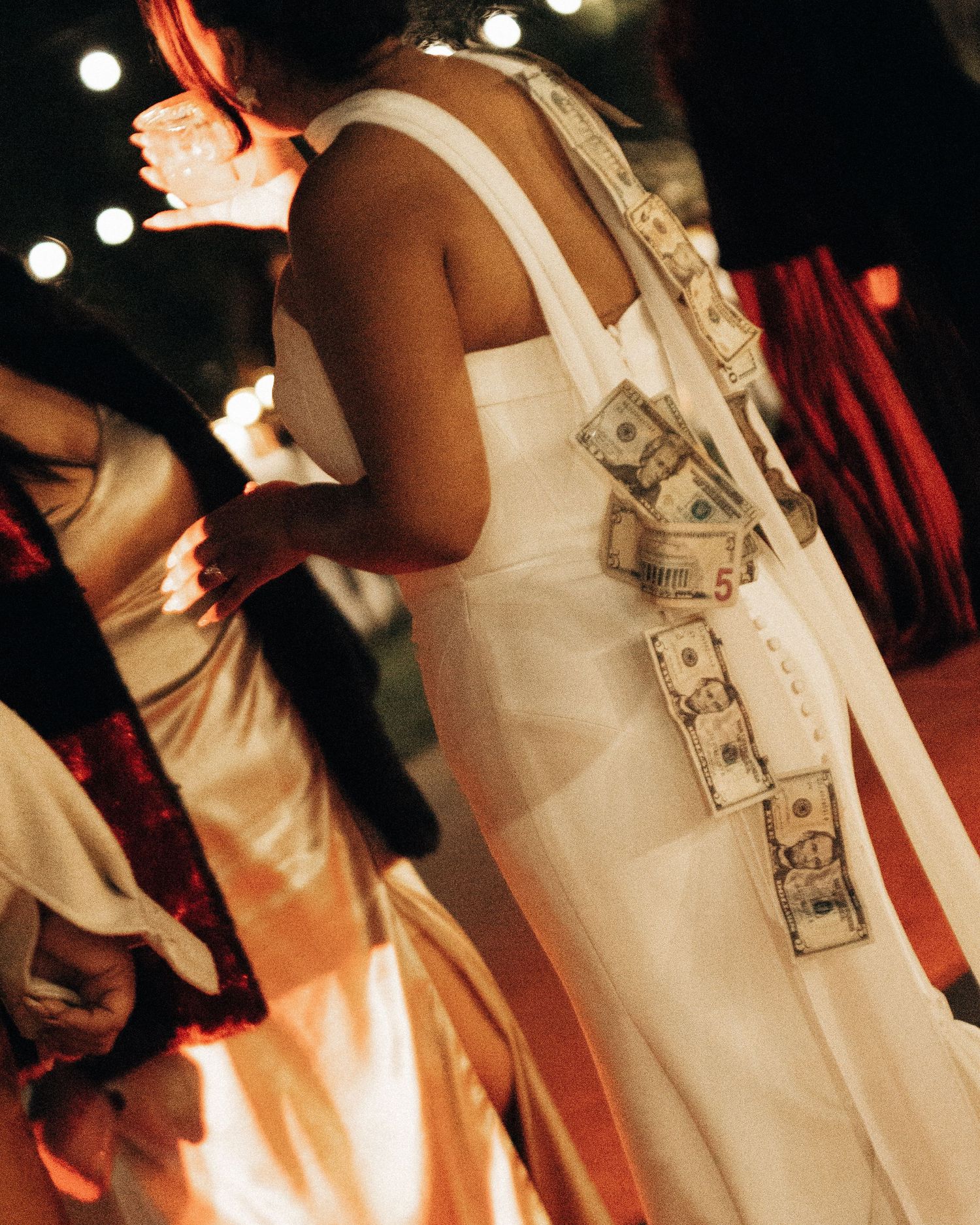 Dollar bills decoratively pinned to a white wedding dress in a dimly lit celebration setting with bokeh lights.
