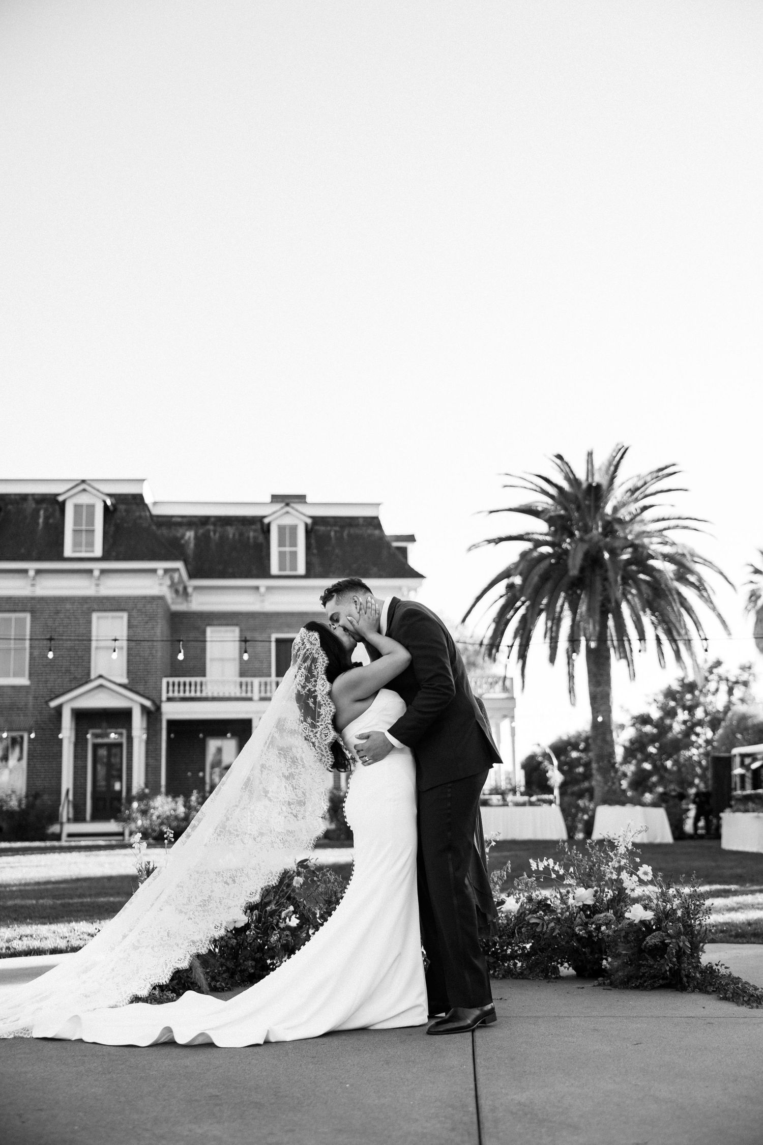 A couple in formal wedding attire pose for romantic photos in front of a historic mansion with palm trees.