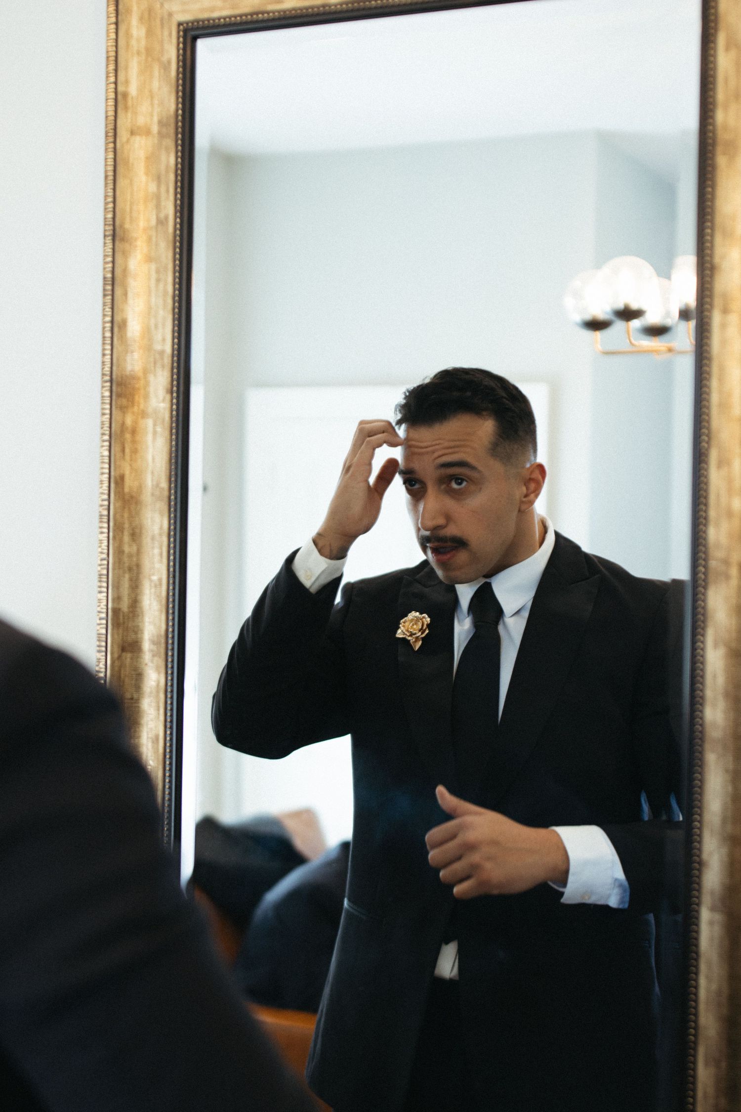 Someone in a black suit and tie adjusts their hair while looking in a mirror with a gold frame.