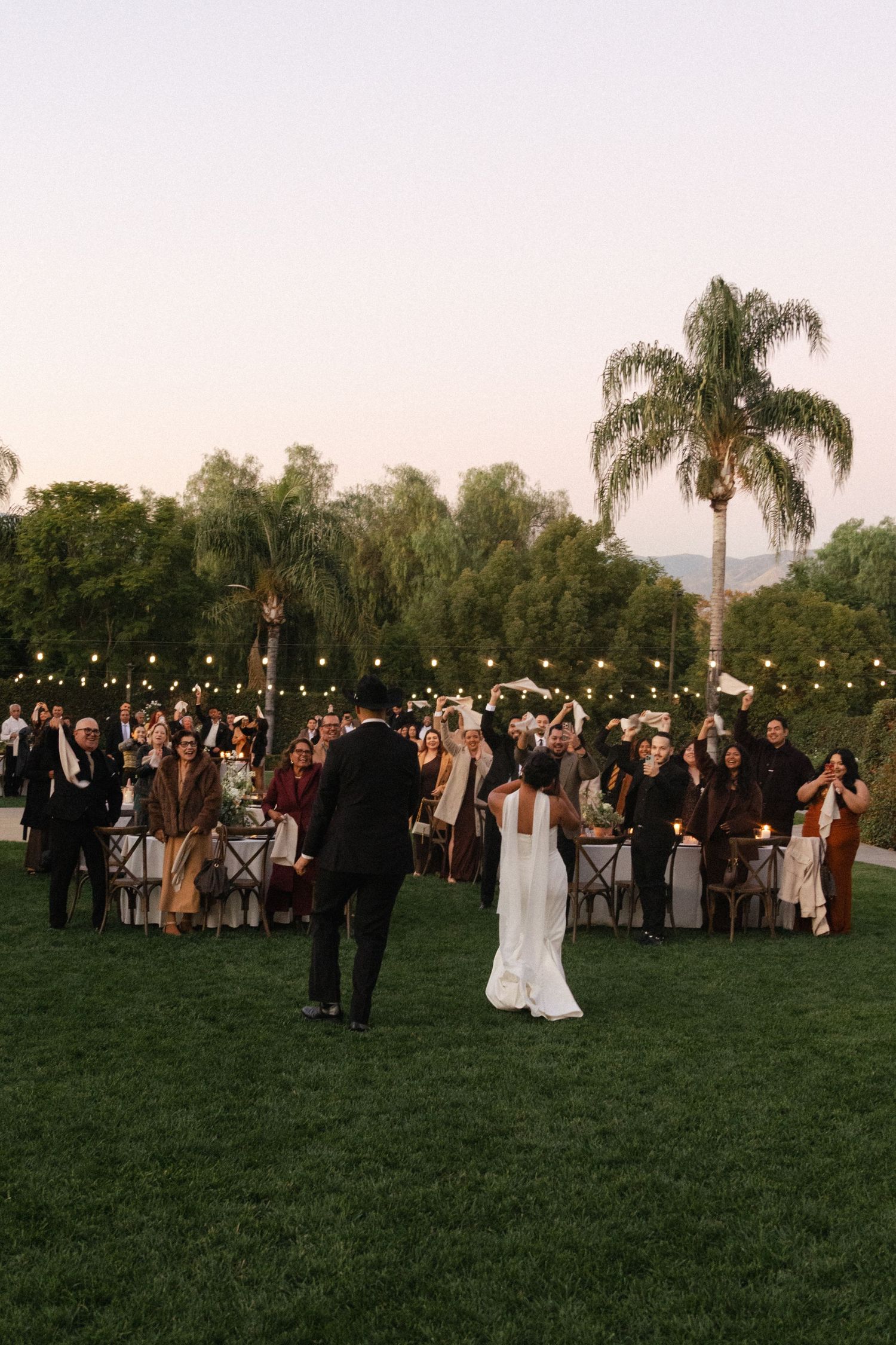 Wedding guests gather on a lush green lawn at dusk with palm trees and string lights creating an elegant outdoor atmosphere.
