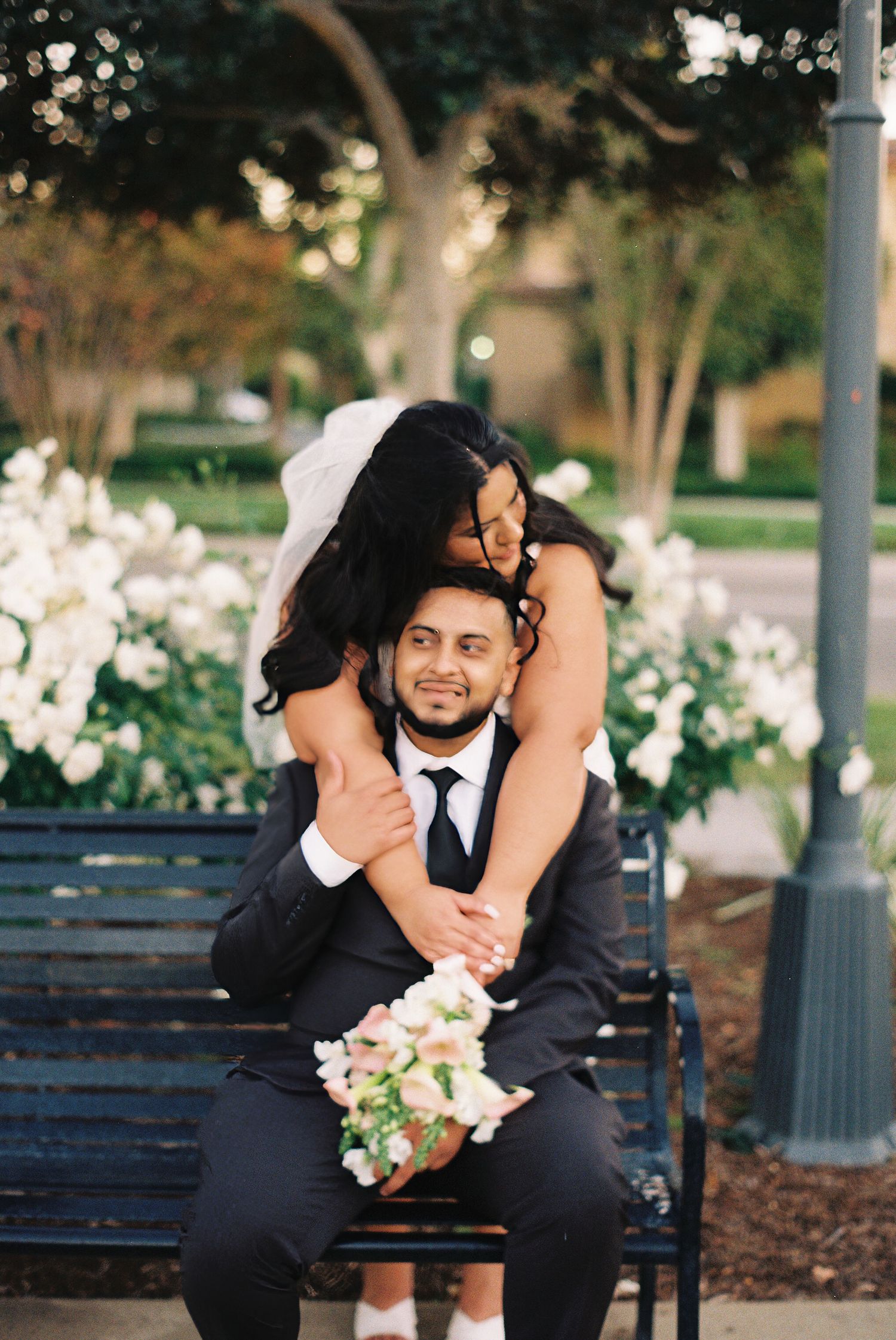 Couple sharing intimate moments on a park bench surrounded by blooming flowers and greenery in an outdoor setting. Micro wedding