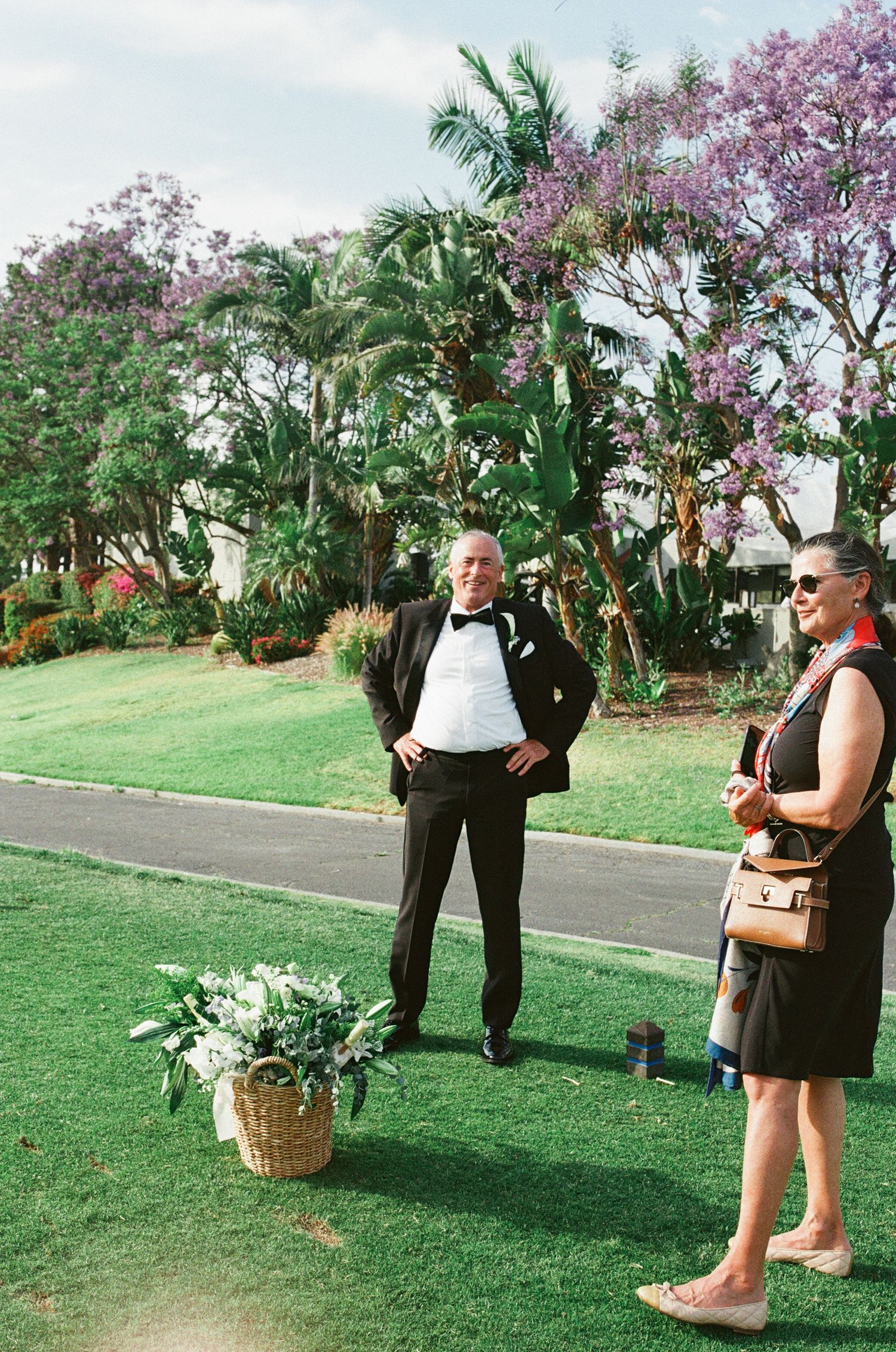 Two people in formal attire stand on a manicured lawn with blooming jacaranda trees and palm trees in the background.