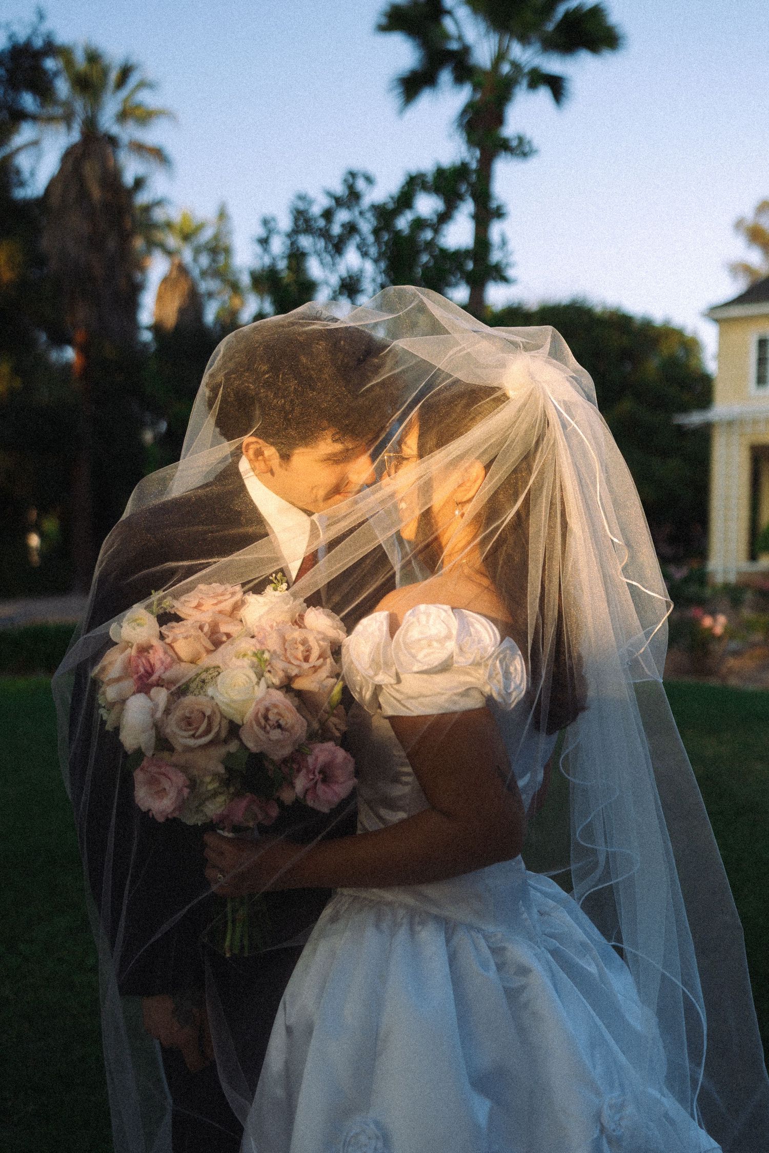 Bride and groom sharing a romantic kiss under a wedding veil, holding a pink rose bouquet at dusk.