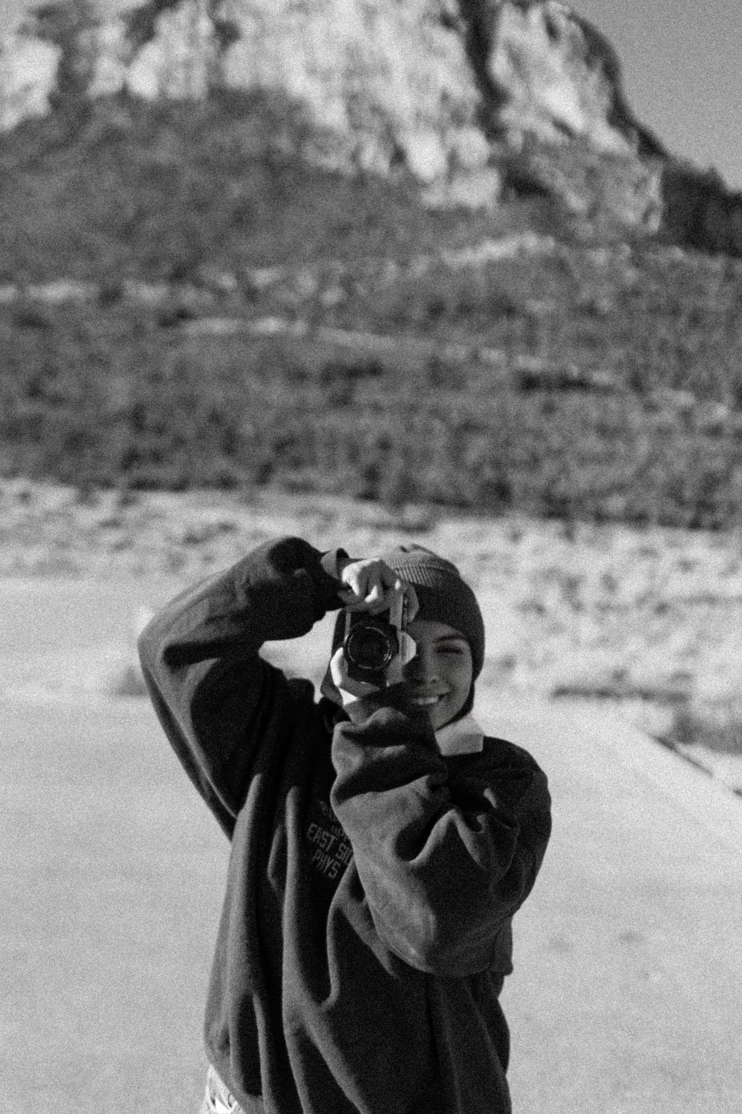 A girl in a dark sweater points a camera towards the viewer with mountains visible in the background. Documentary photographer