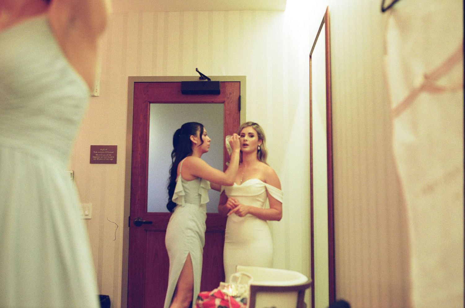 Two people in elegant attire preparing and adjusting outfits in a brightly lit dressing room with a magenta door.