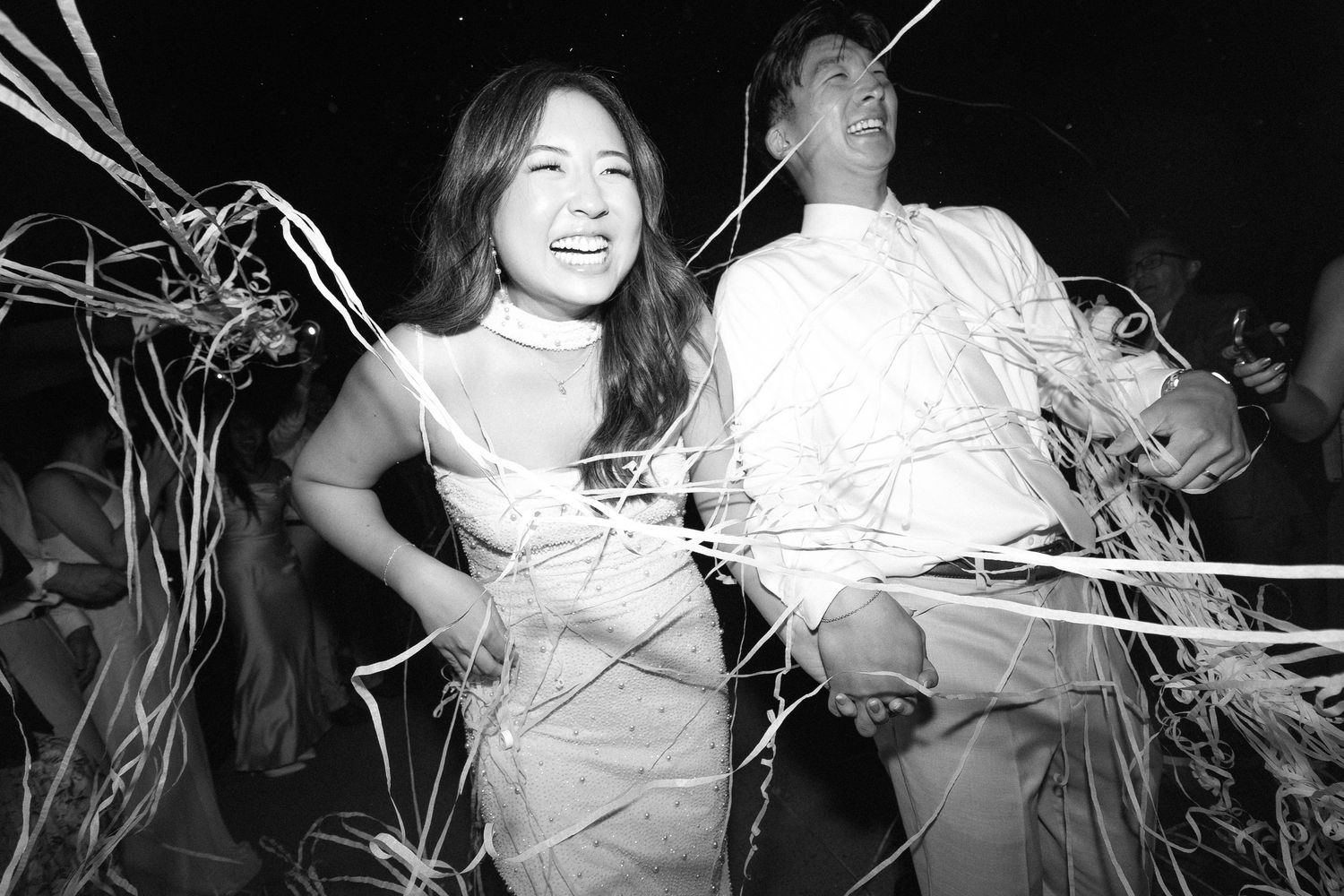 Black and white photo of a laughing couple covered in streamers at a festive celebration or wedding party.