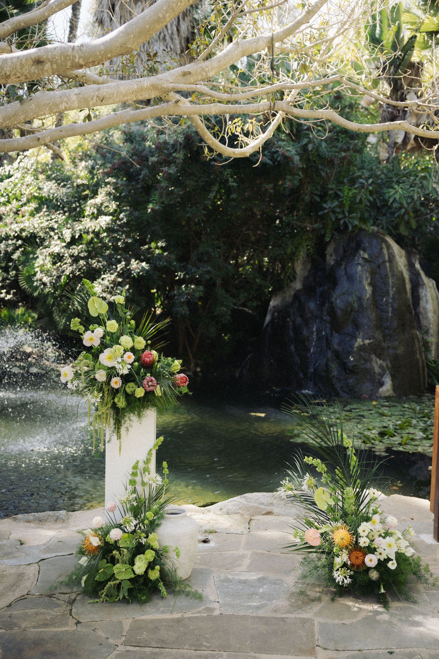 Outdoor wedding ceremony altar with white floral arrangements, tropical greenery, and a serene pond backdrop.
