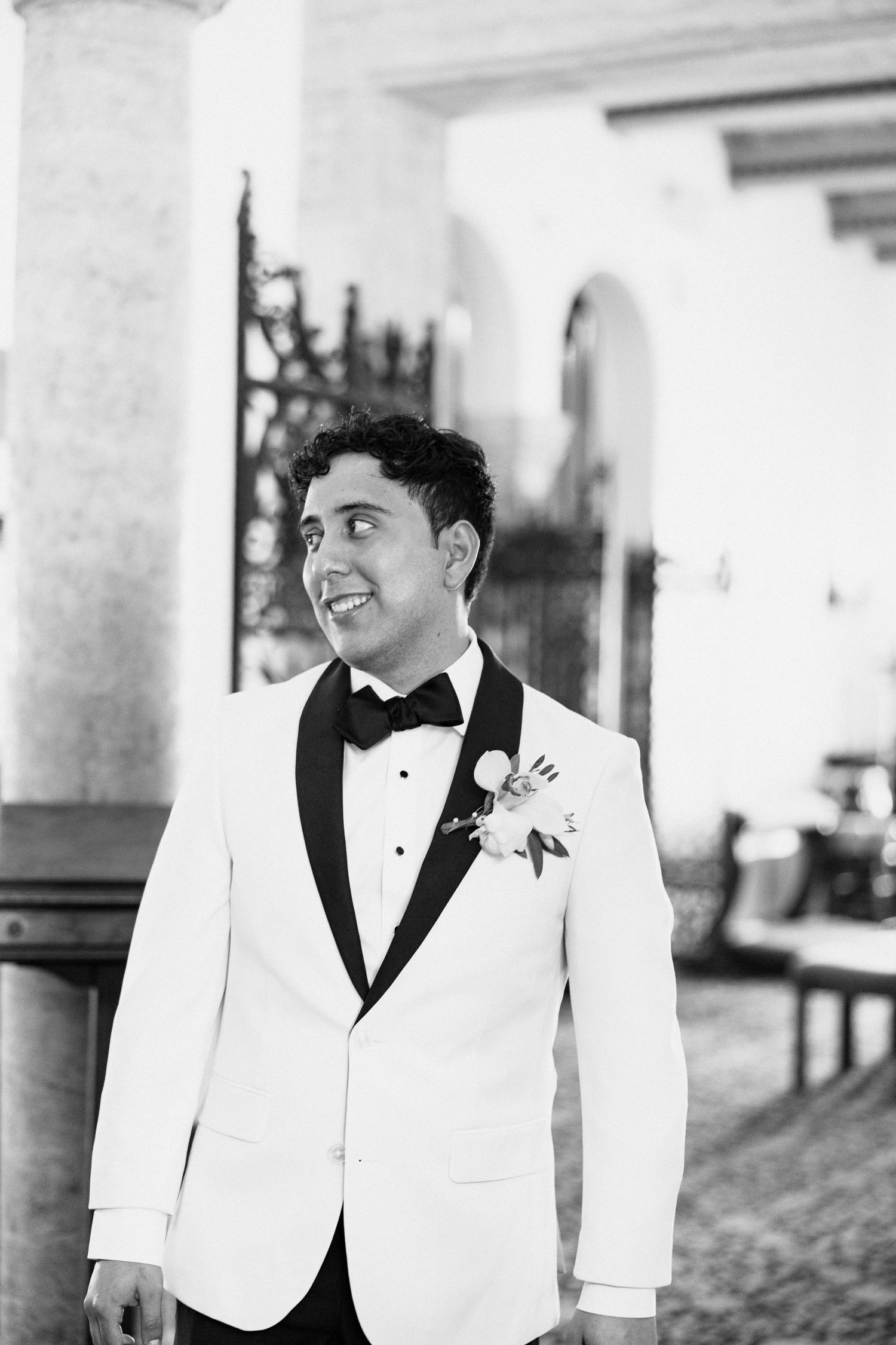 A person in a white tuxedo jacket and black bow tie smiles while looking away from the camera during a formal event.