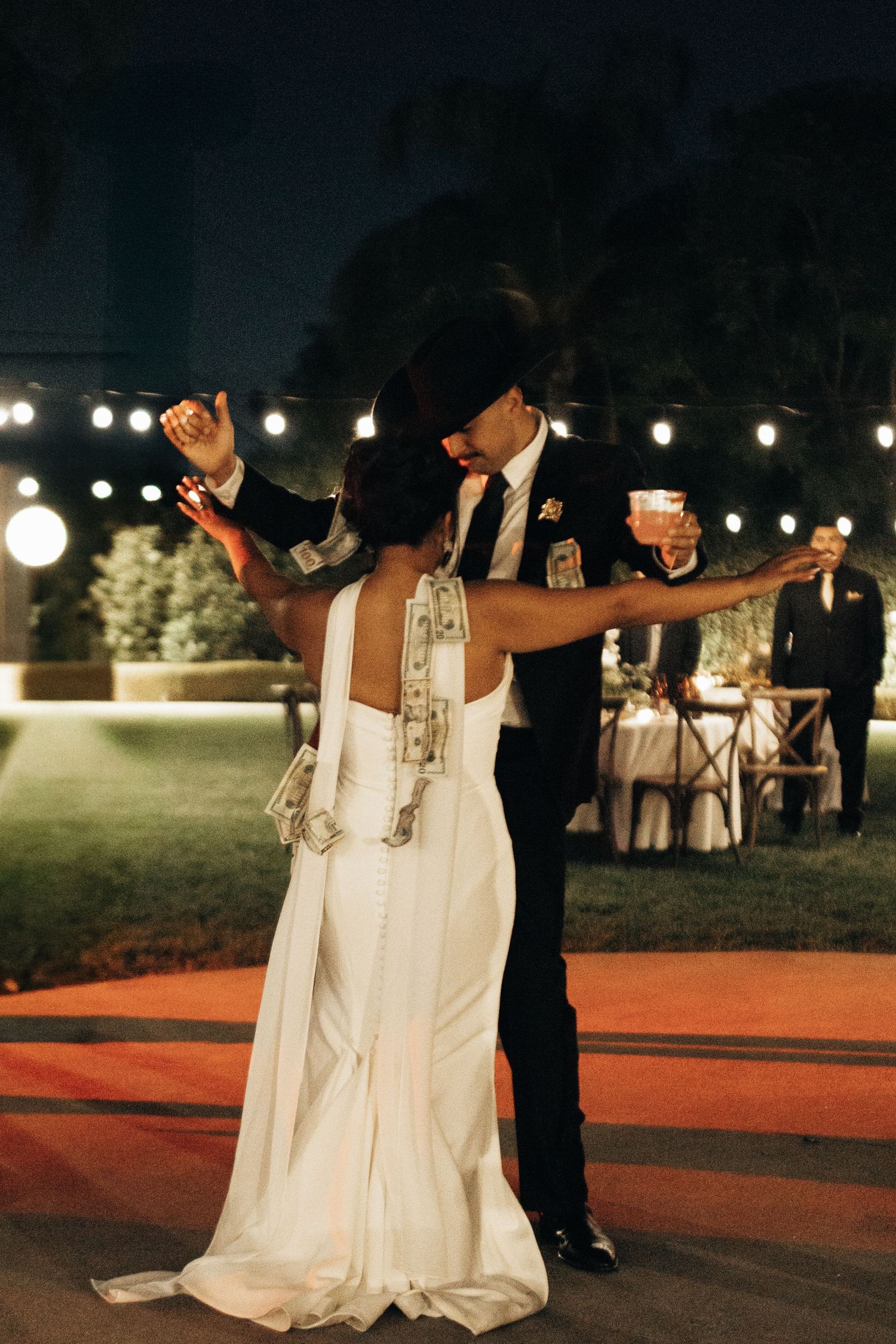 Couple shares their first dance at outdoor evening wedding reception with string lights and glowing lanterns in background.