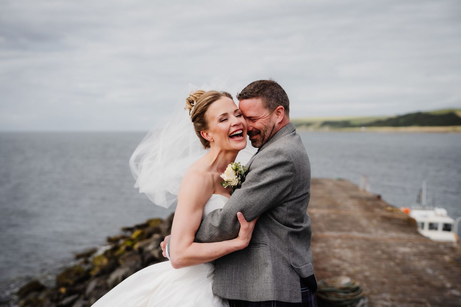 Happy bride and groom embracing on a seaside pier, with veil blowing in the wind on a cloudy day.